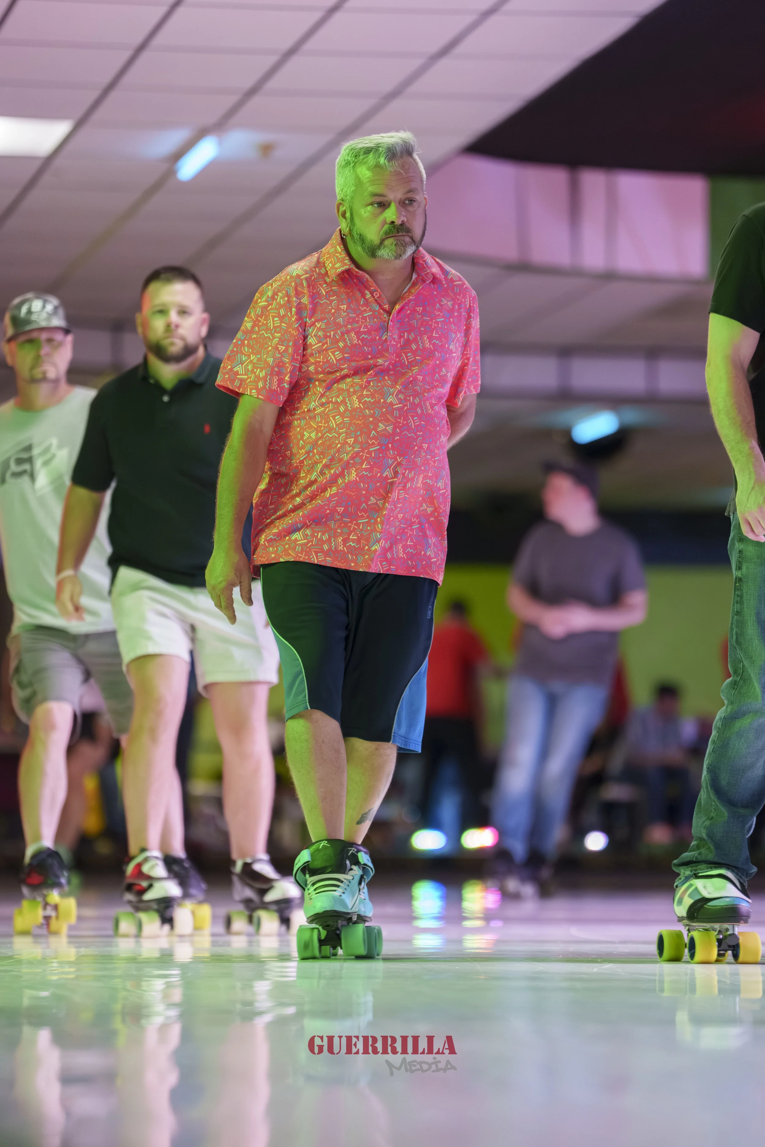 A group of people roller-skating indoors, with a man in a colorful pink and orange shirt leading the group, and other skaters visible in the background.