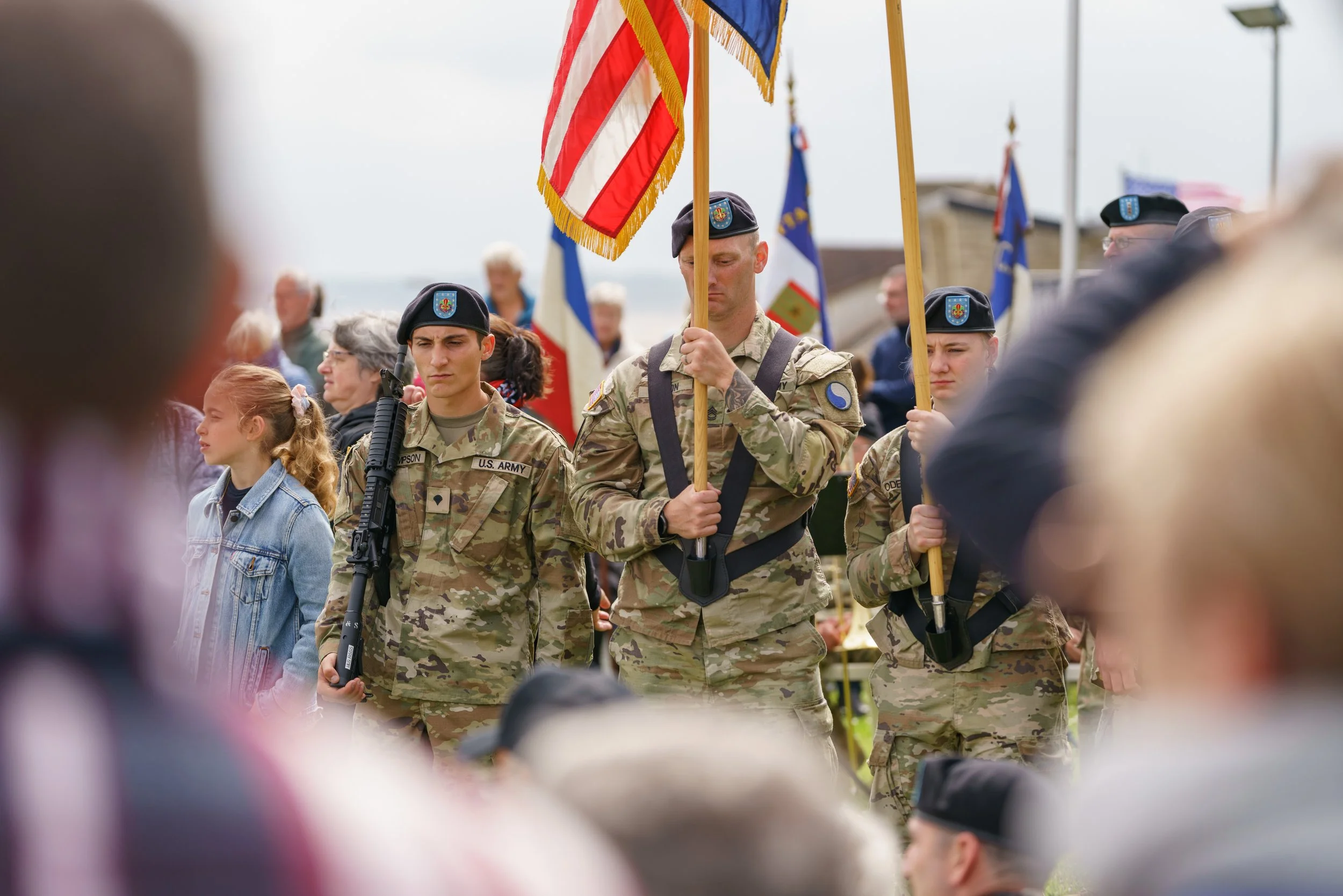 U.S. Army soldiers in camouflage uniforms participating in a ceremony, standing at attention with flags, with a crowd observing in the background.