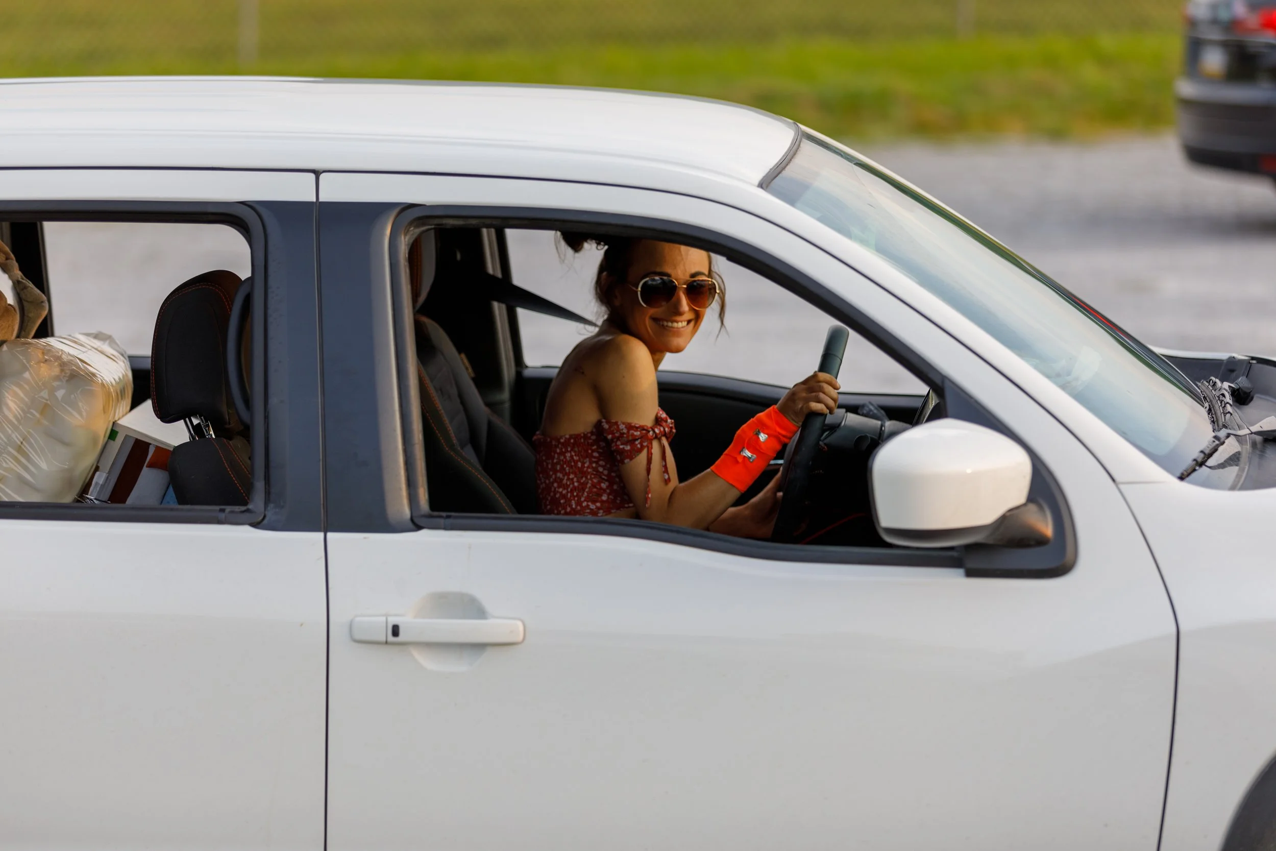 Woman sitting in the driver's seat of a white car, smiling, wearing sunglasses, an off-shoulder red floral dress, and orange gloves, holding the steering wheel.