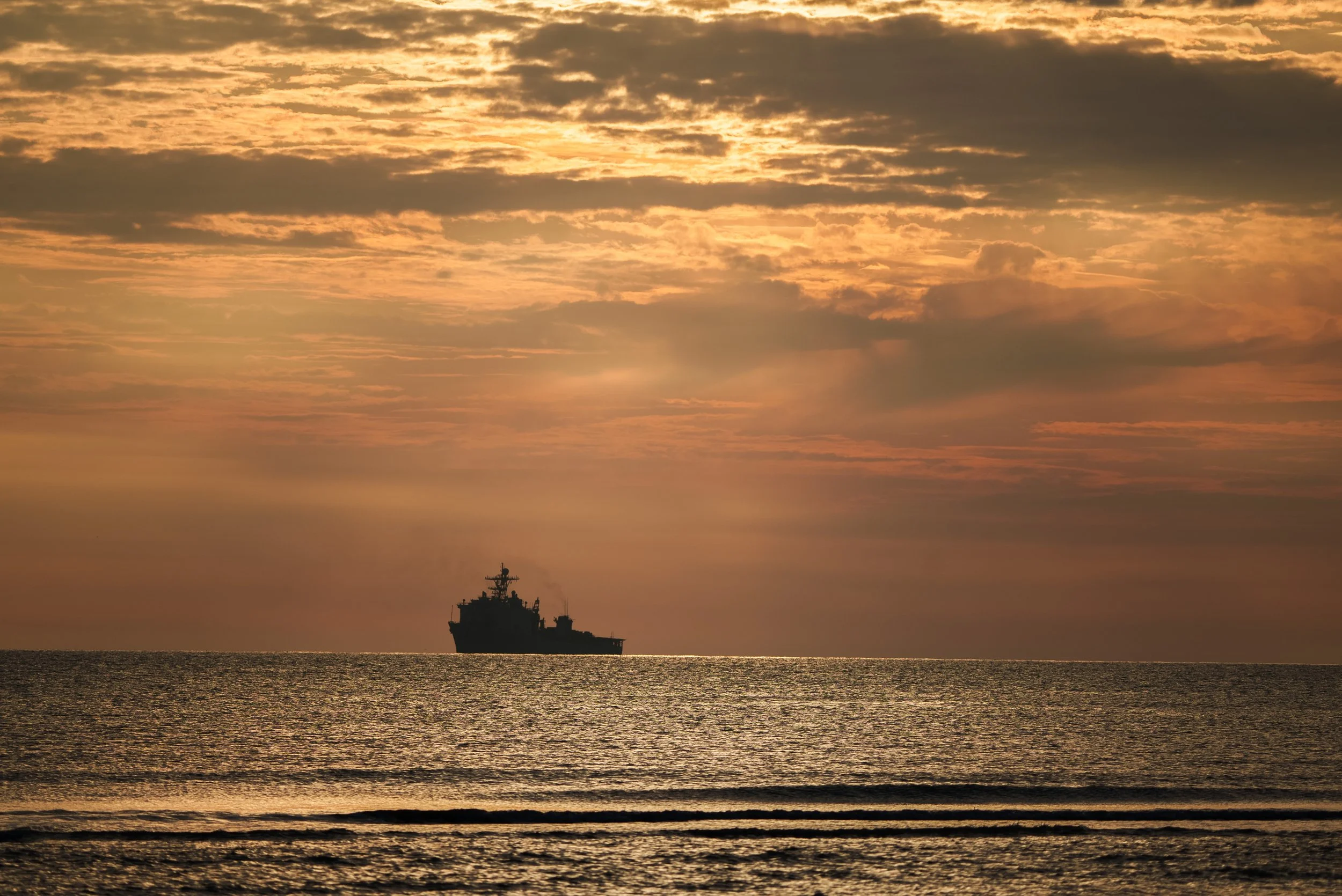 A silhouette of a battleship on calm ocean waters during a sunset with a partly cloudy sky.
