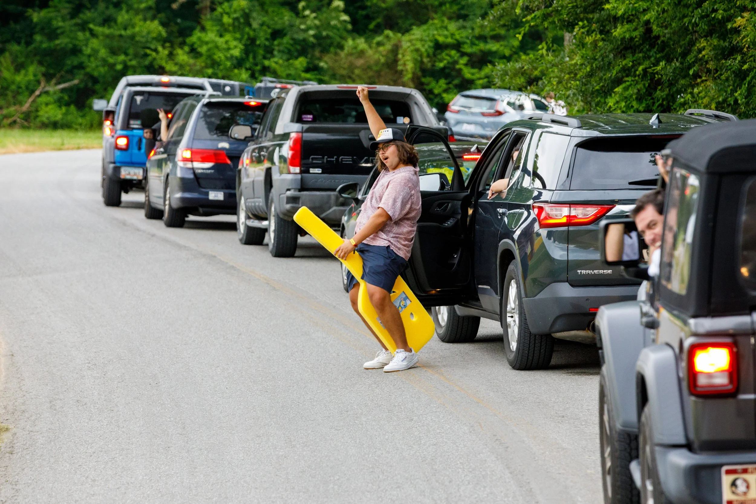 People in a line of cars on a country road, including a woman holding a yellow float, with trees in the background.