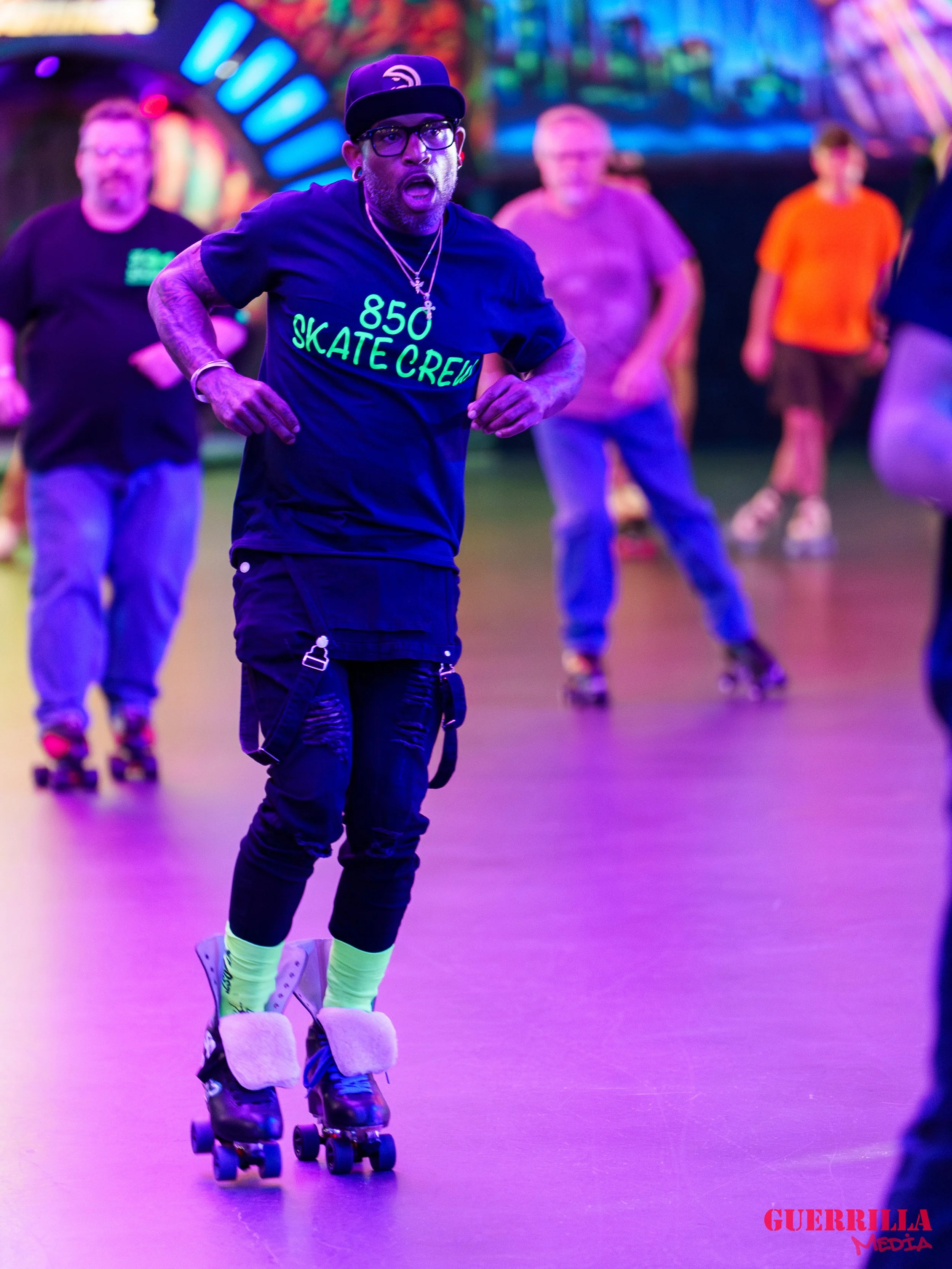 A man wearing glasses, a black cap, a black T-shirt with neon green writing, ripped black pants, neon green socks, and roller skates, is skating in an indoor roller rink with multicolored lighting. Several other people are skating in the background.