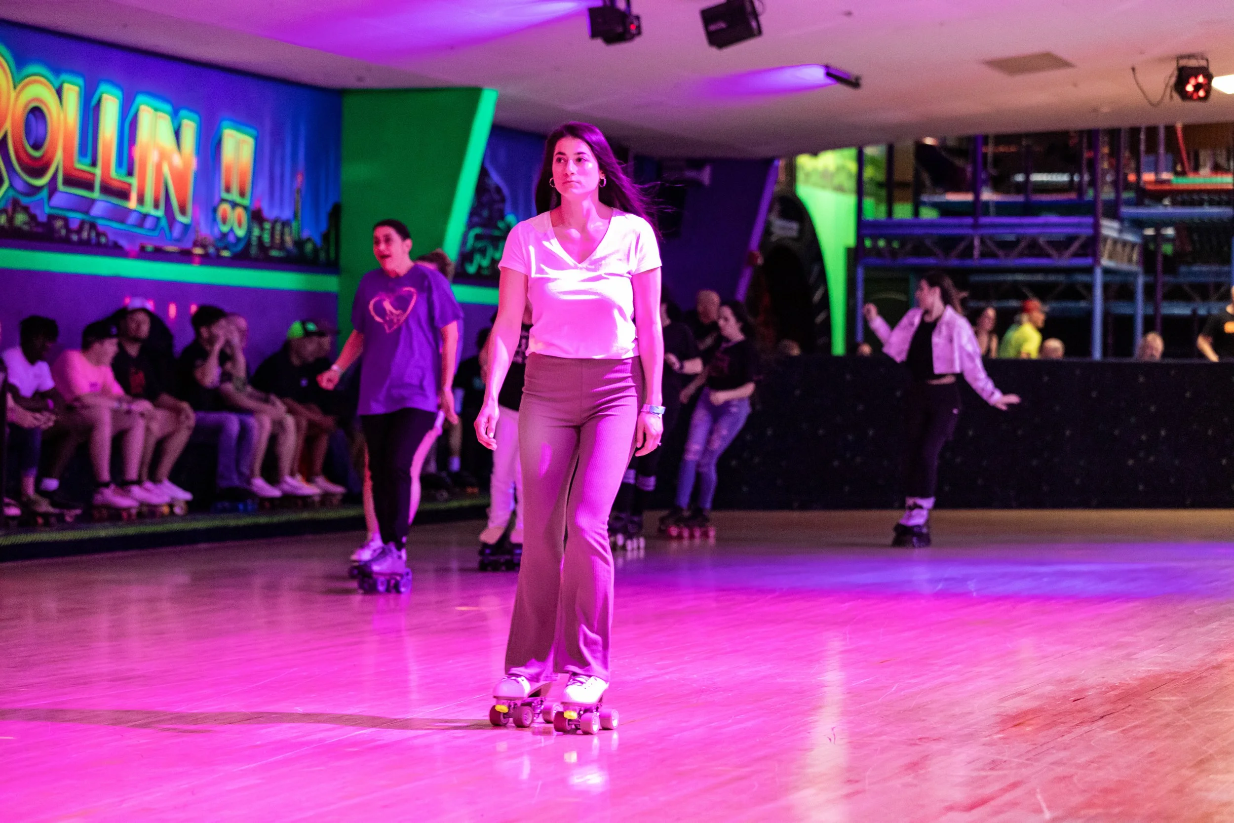 People roller skating in an indoor roller rink with neon lighting, with a woman in a white top and beige pants in the foreground, and others sitting on benches and skating in the background.