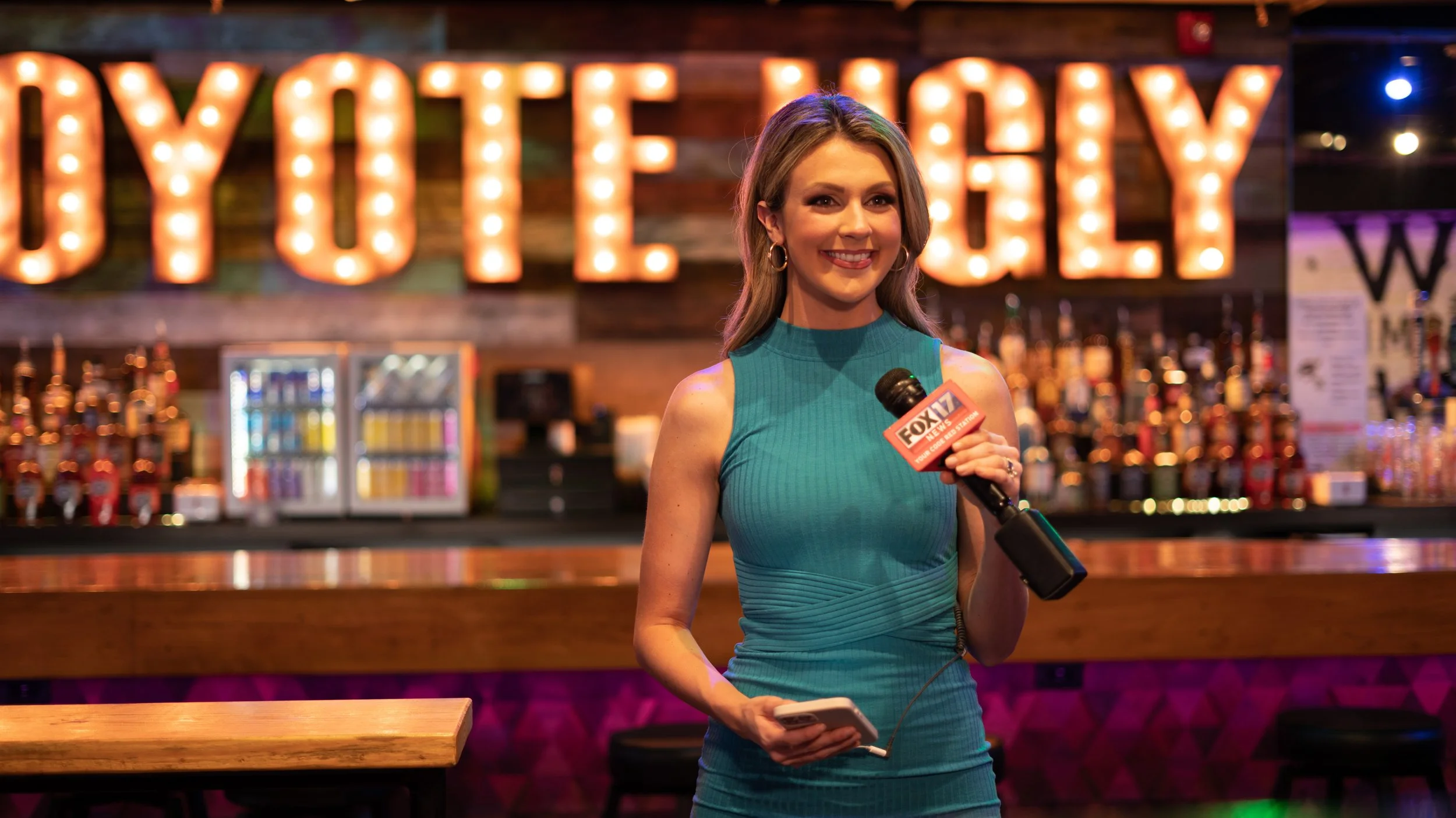 Female reporter in teal dress holding a microphone with a "FOX 17" logo, standing inside a bar or restaurant with a "Lounge" sign in lights in the background.