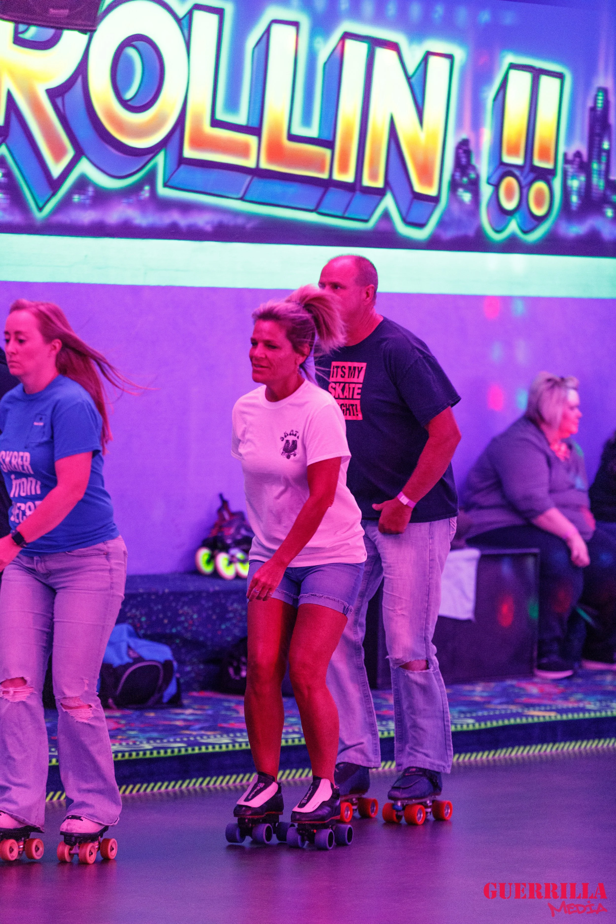 People roller skating in an indoor rink with neon lights and a colorful 'Rollin'! sign in the background.