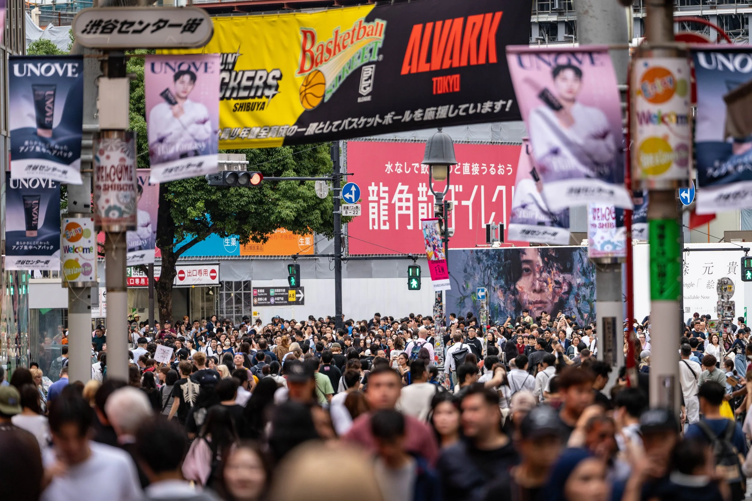 A bustling street in Shibuya, Tokyo filled with a large crowd of pedestrians. There are various colorful advertisements and banners hanging overhead, including a prominent basketball-themed banner and signs in Japanese. Traffic lights and trees are v