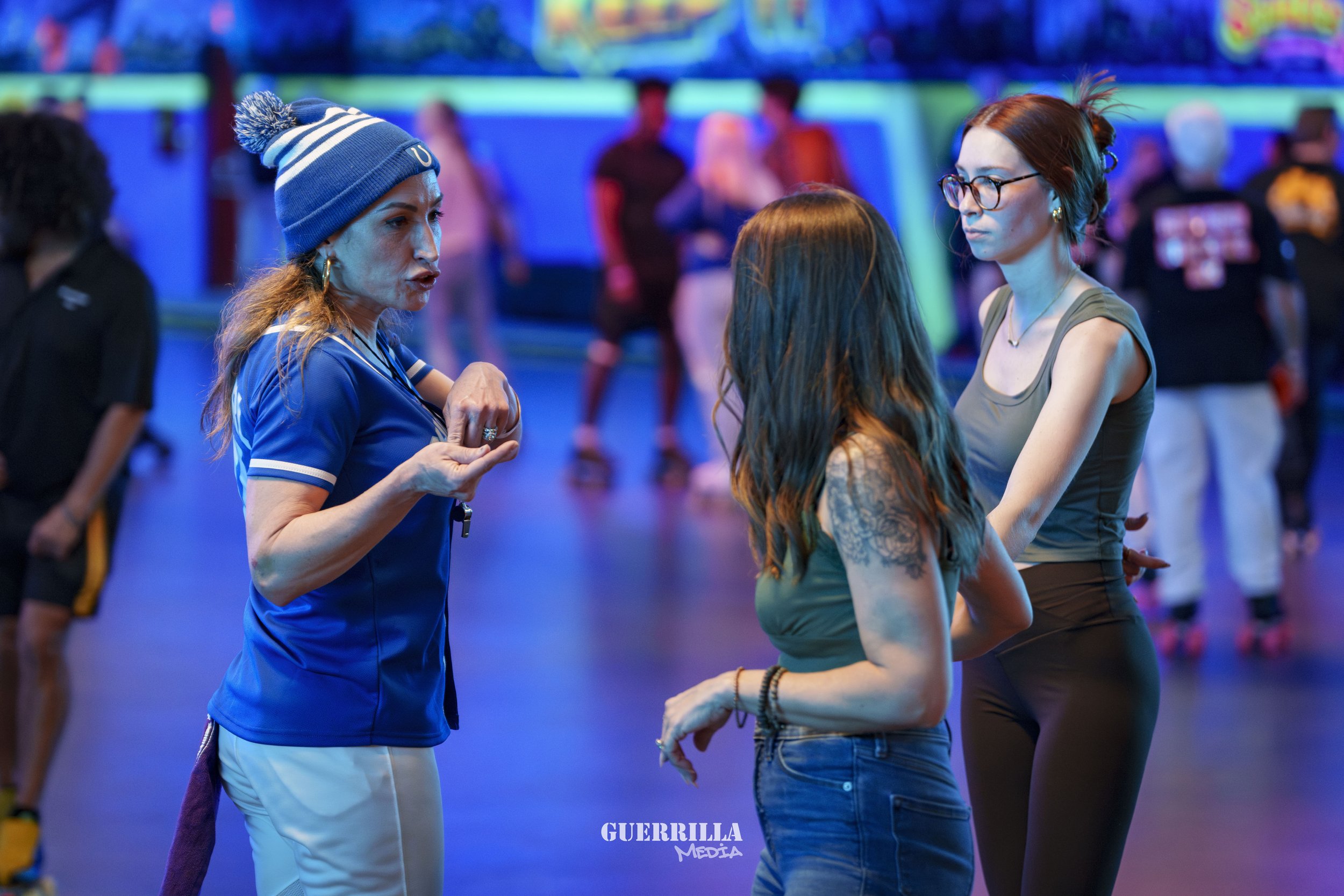 Three women are having a conversation in a brightly lit indoor skating rink, with roller skaters and colorful lights in the background.