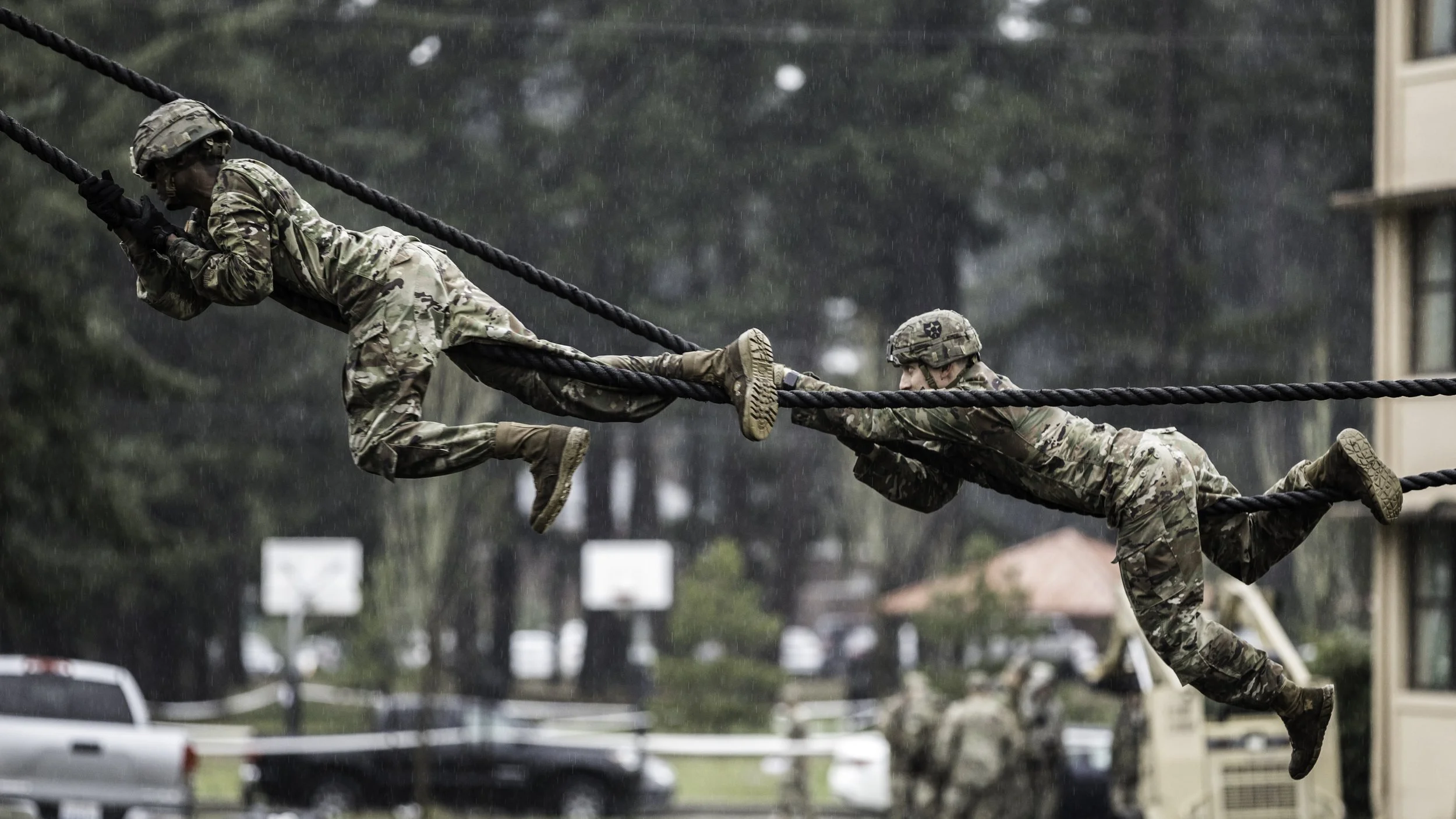 Two soldiers in camouflage uniform and helmets are crossing a rope obstacle during a rainstorm, each holding onto a thick black rope and using their legs to climb.