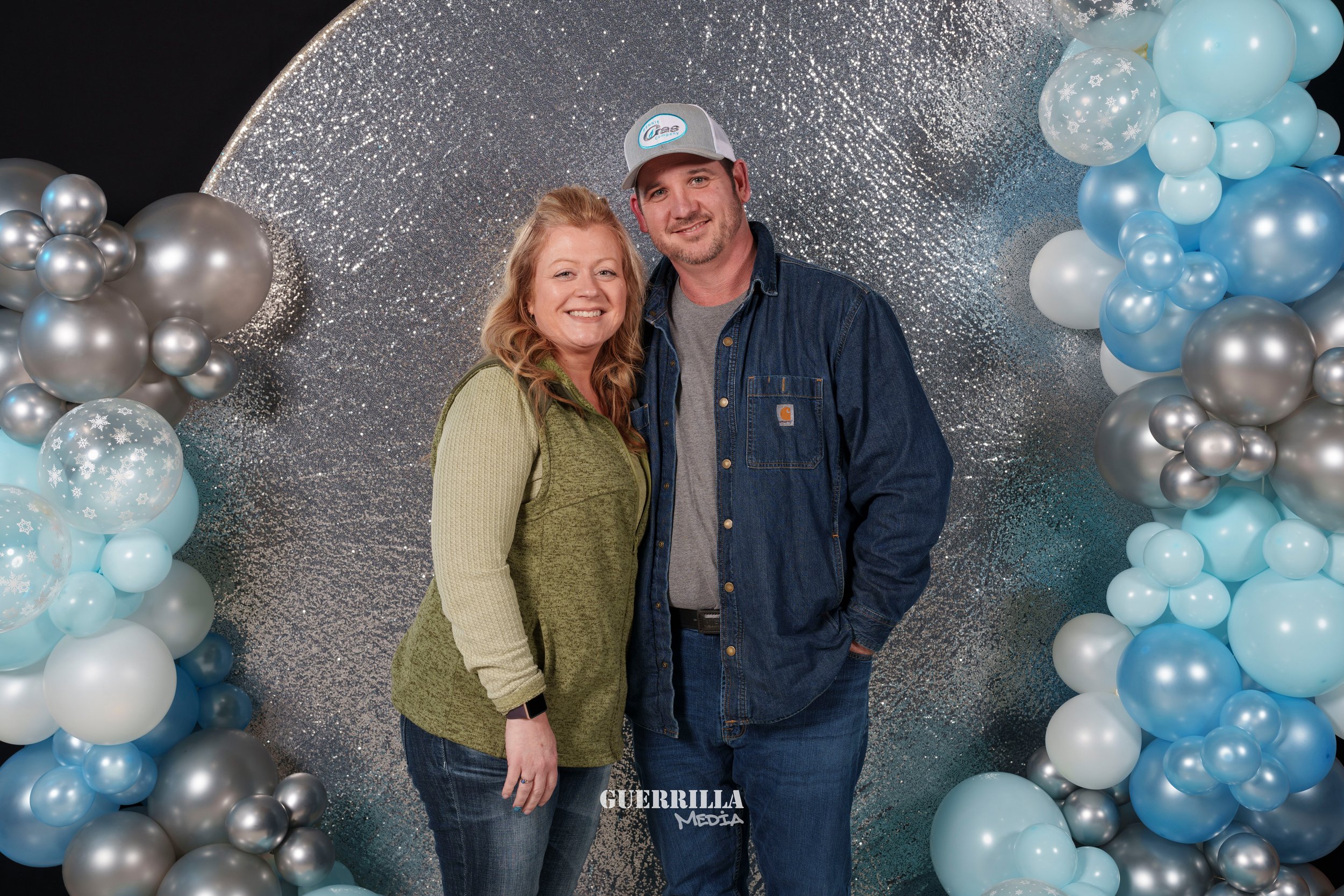 Smiling man and woman posing in front of a silver and blue balloon arch and a glittery backdrop.