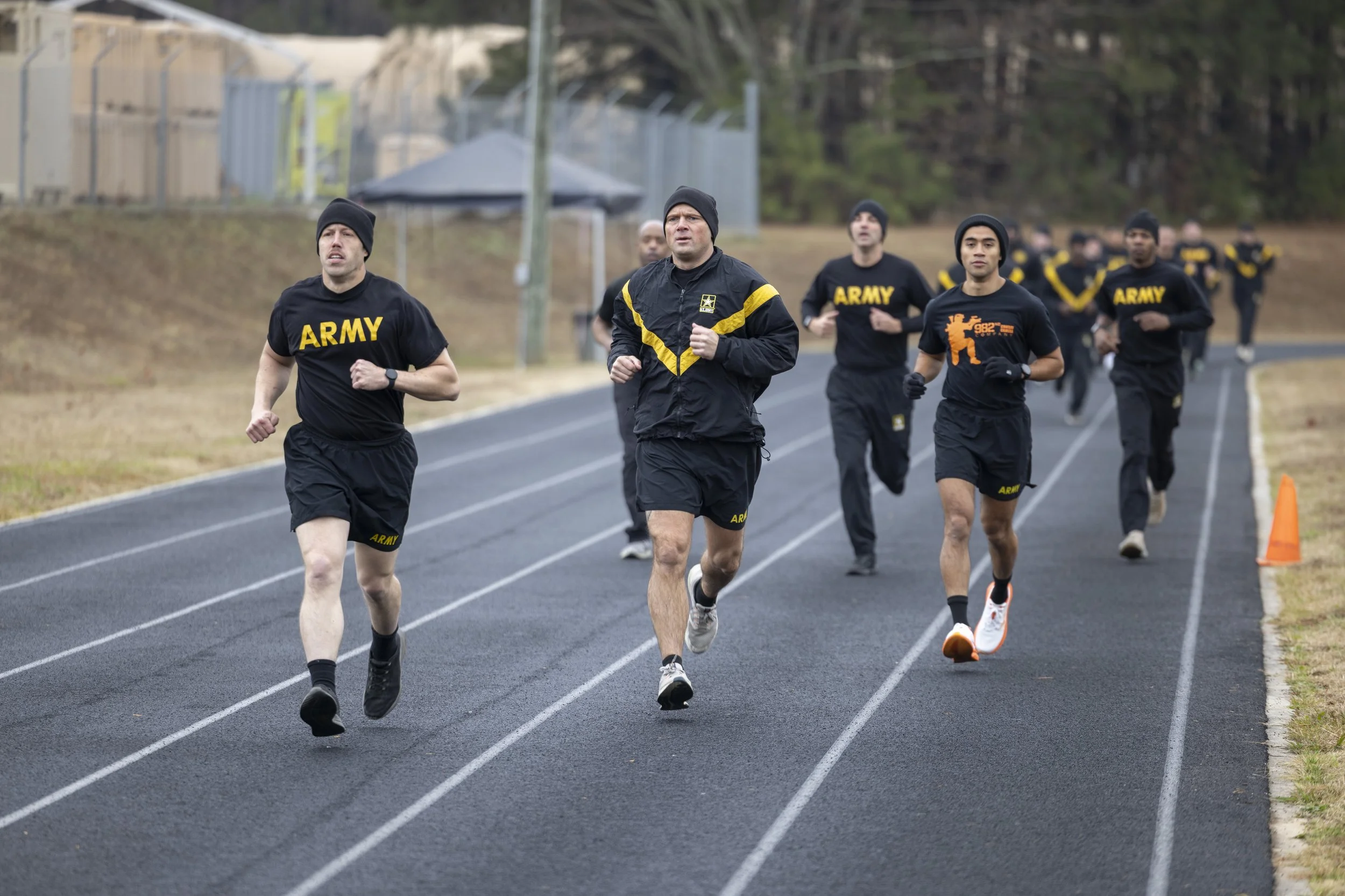 Group of men running on a track, wearing athletic clothes with 'ARMY' printed on their shirts, outdoors during daytime.