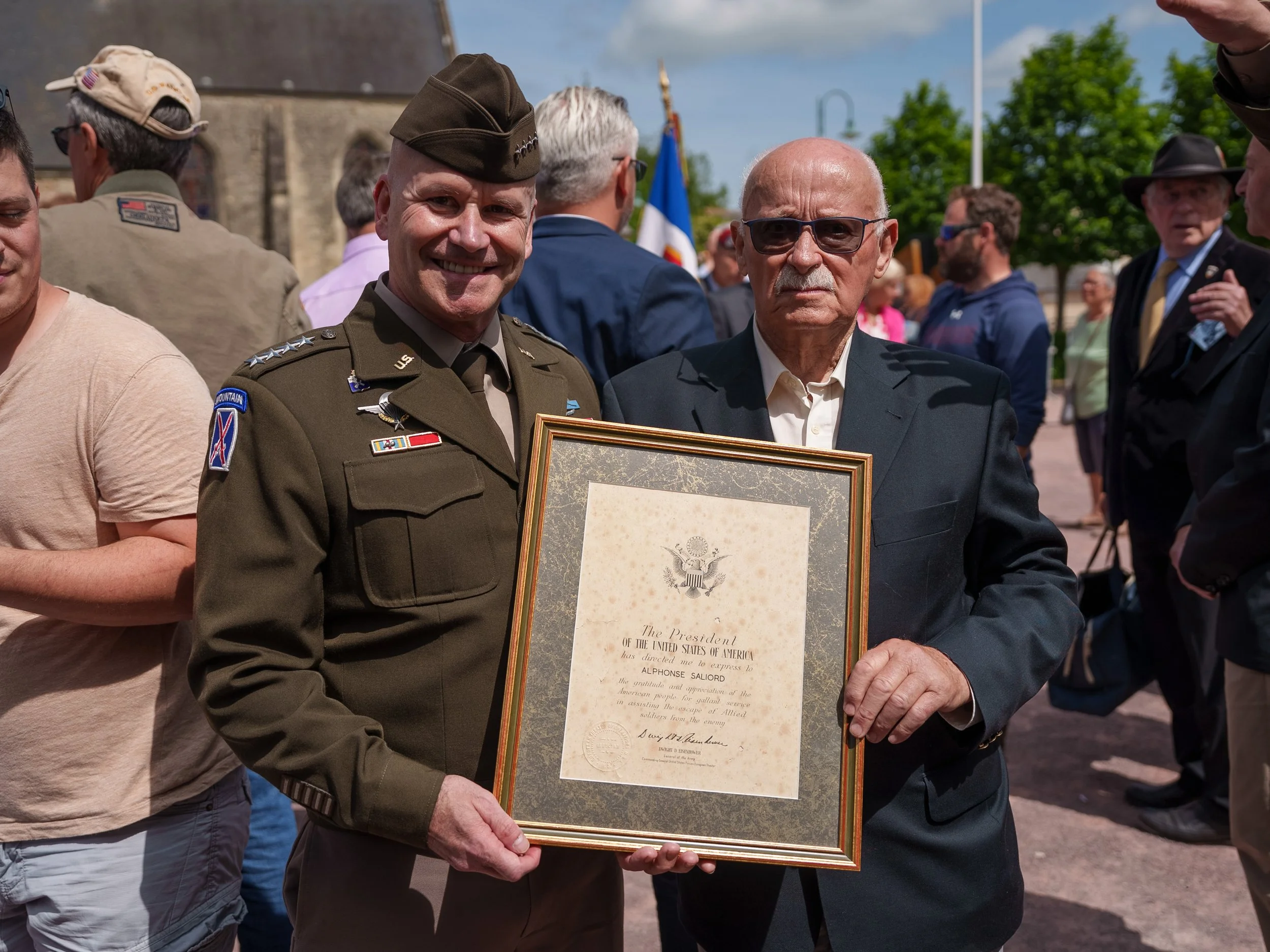 Two men, one in a military uniform and the other in a suit, holding a framed certificate at an outdoor event with a crowd in the background.