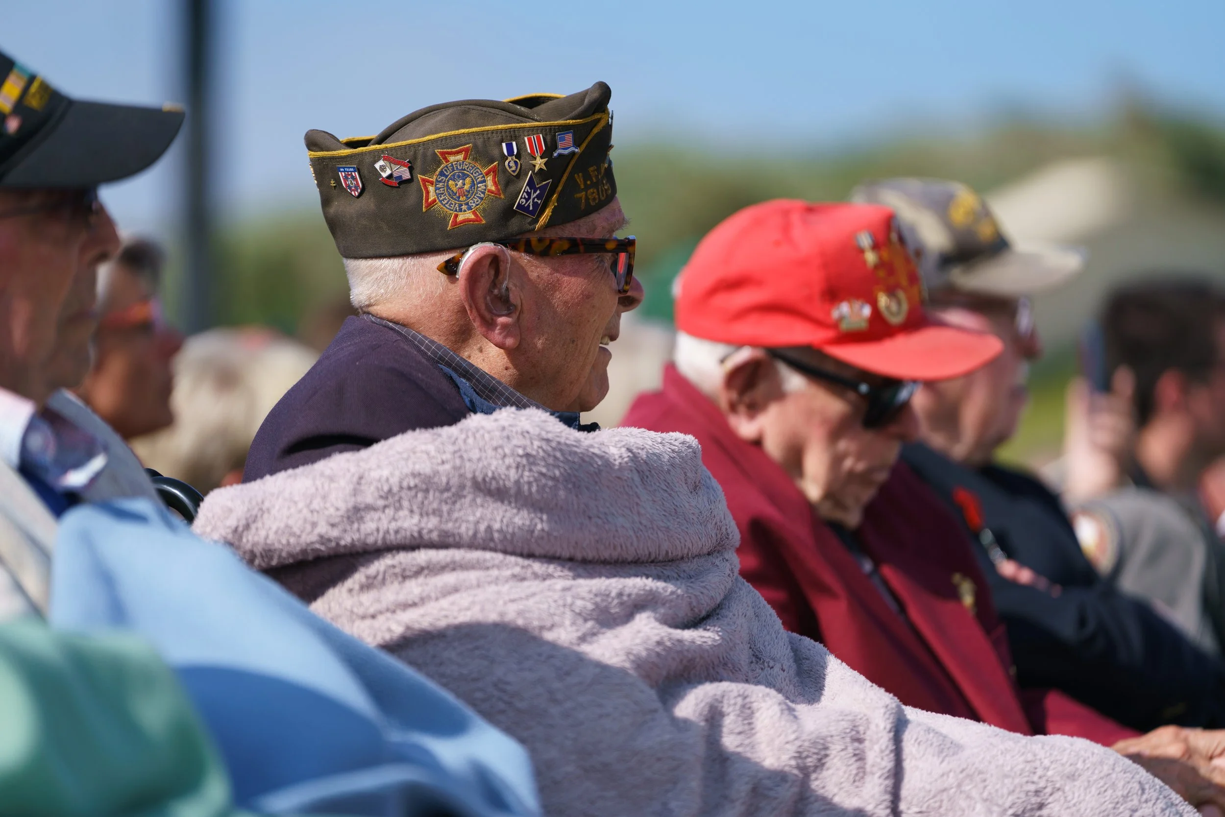Group of senior veterans sitting outdoors during daytime, wearing hats with military patches and medals, some with sunglasses, in a commemorative or memorial event.