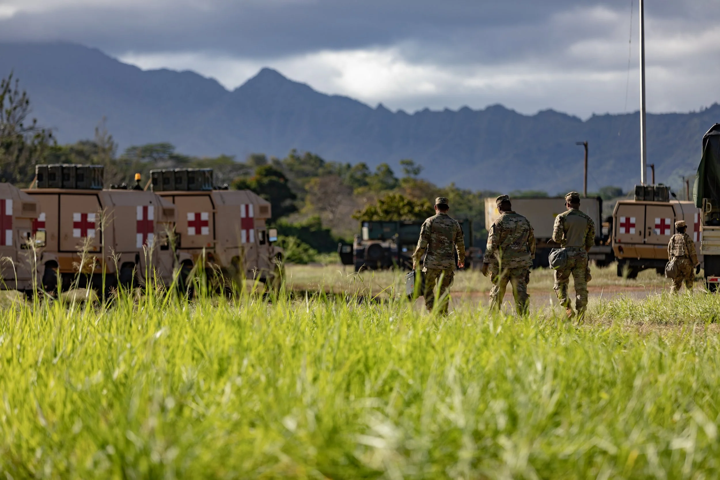 Military personnel walking in a grassy field with supply trucks bearing red cross symbols, mountains in the background, and cloudy sky above.