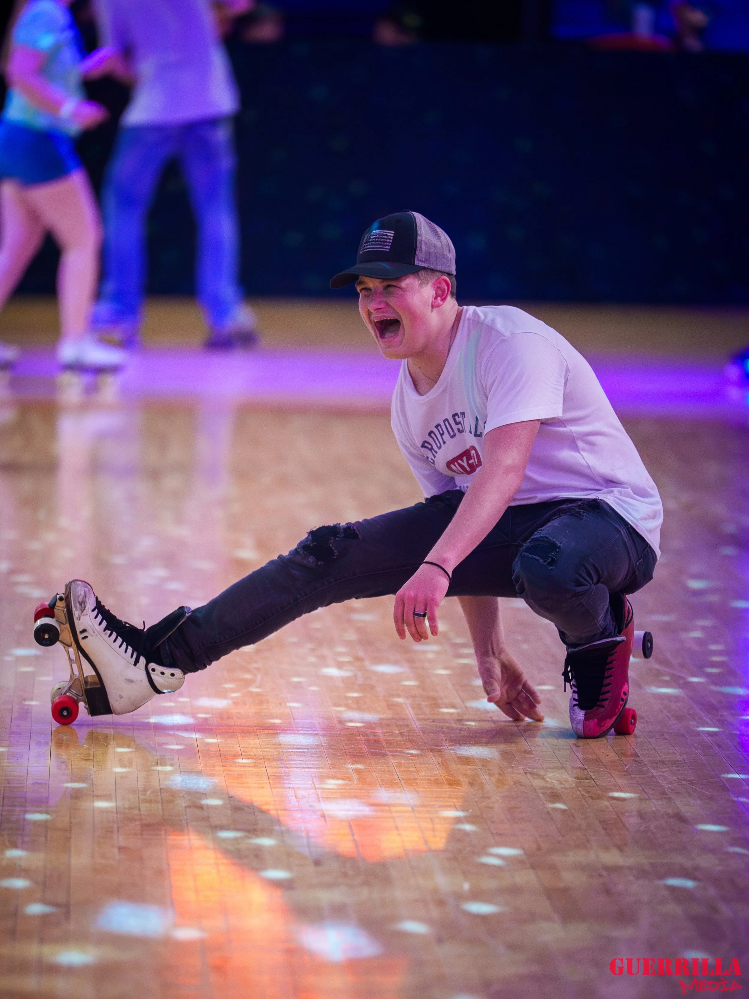 Person in white t-shirt and black ripped jeans roller skating on a wooden floor, crouched with arms on the ground, laughing. Two other people skating are blurred in the background.