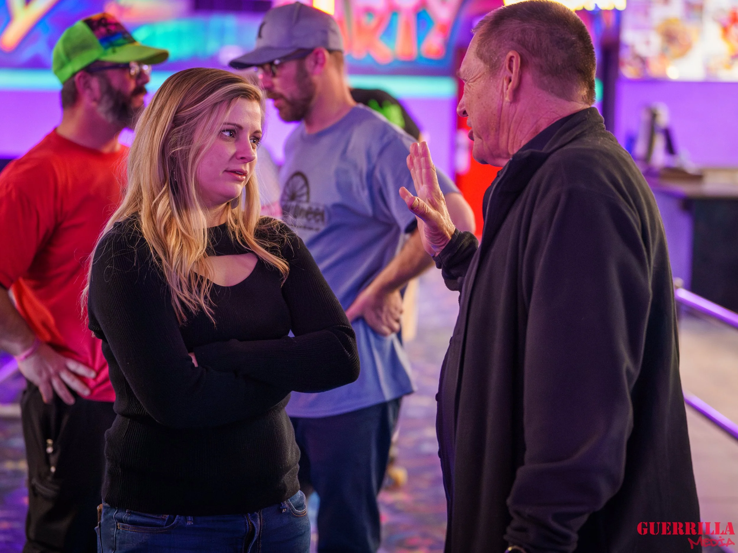 A young woman with blonde hair, wearing a black sweater, appears upset as she listens to an older man with short hair, wearing a dark jacket, who is speaking to her with his hand raised in a stopping gesture. They are in a colorful indoor setting wit