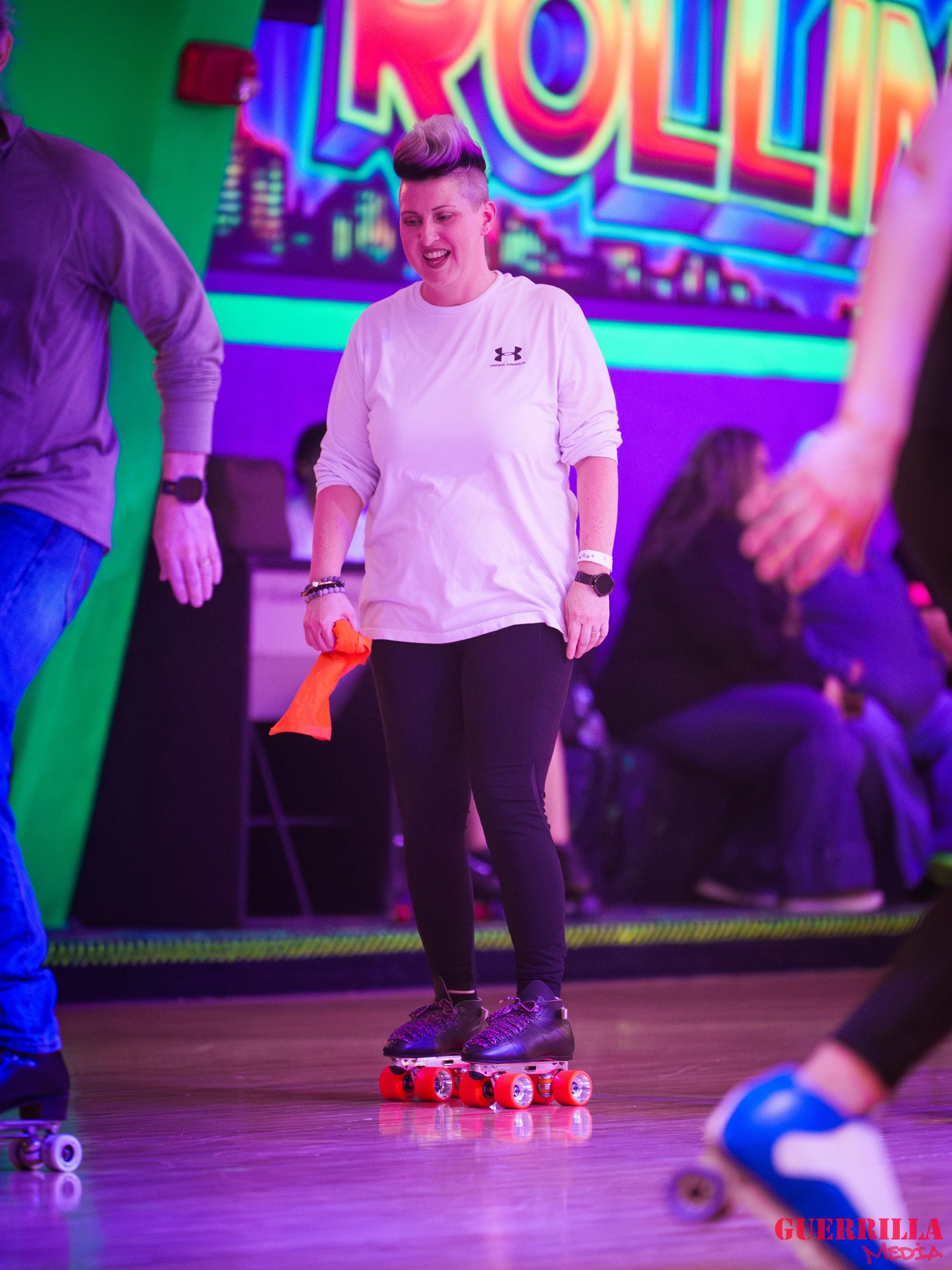 A woman roller skating indoors with a neon sign in the background, holding an orange cloth, wearing a white long-sleeve shirt, black pants, and black roller skates with red wheels.