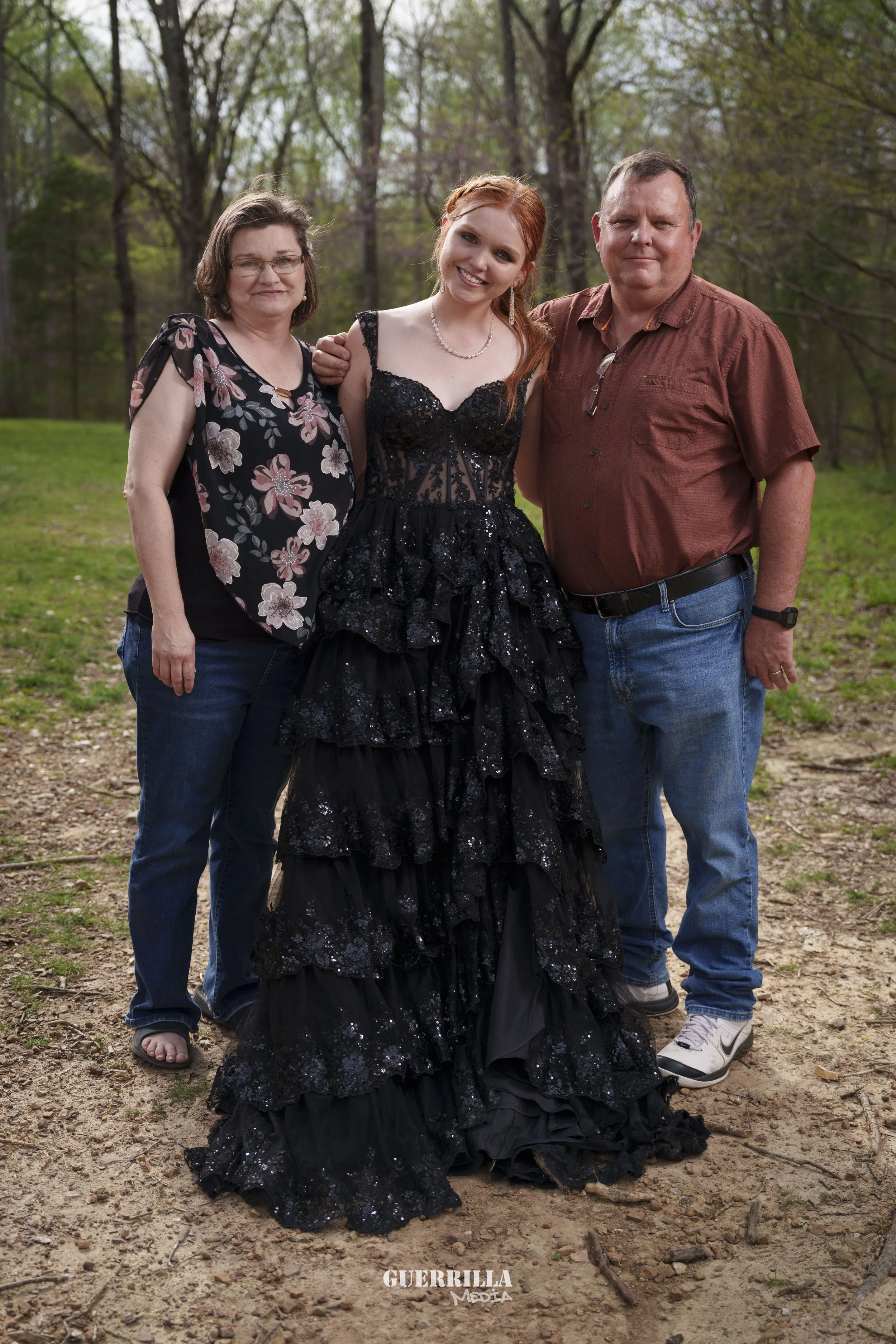 A young woman in a black, ruffled, sequined gown stands between an older woman and older man outdoors in a wooded area.