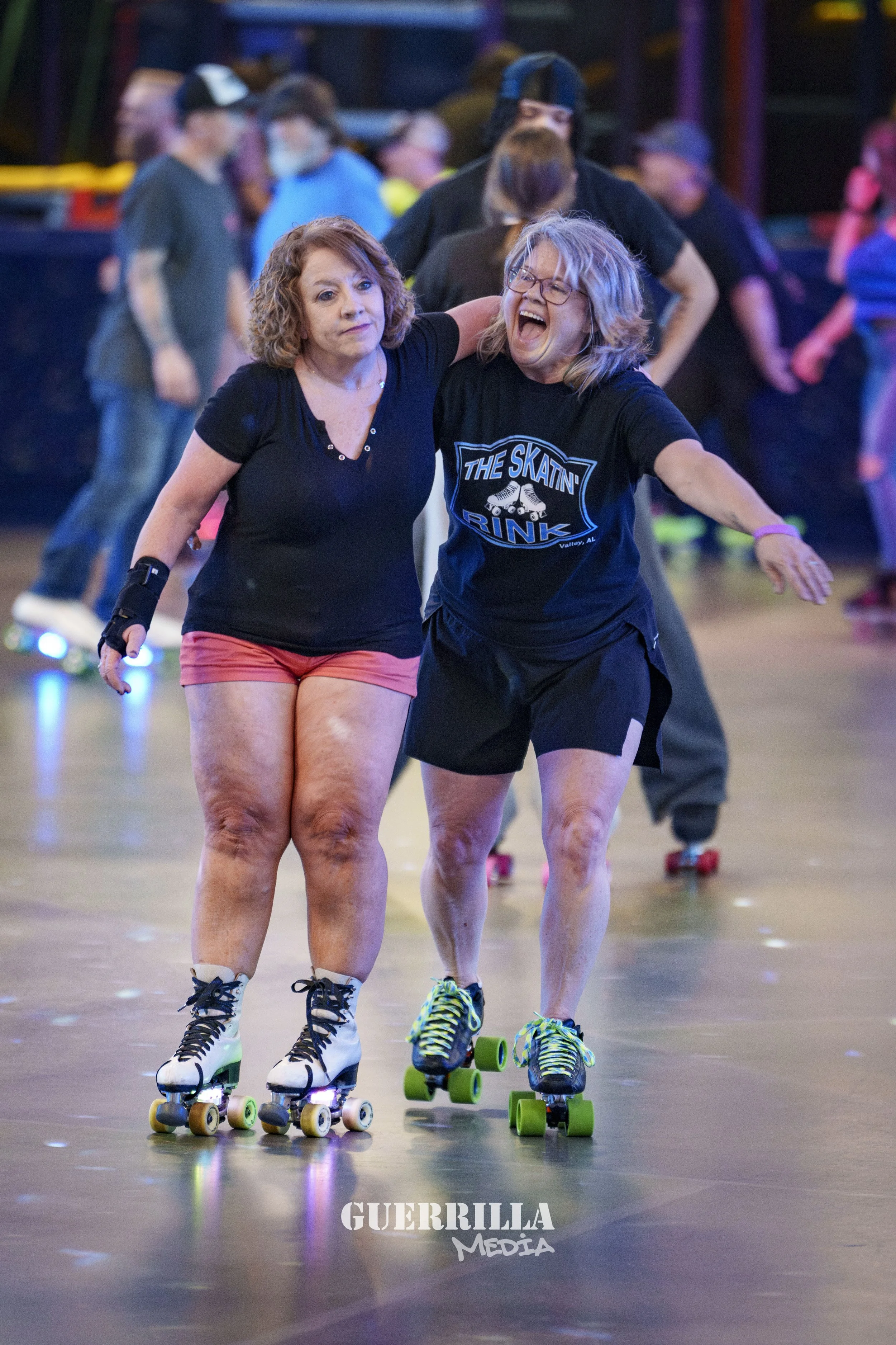 Two women roller skating indoors, one with her arm around the other's shoulder, both smiling and enjoying the moment. The background shows other skaters and colorful lighting.