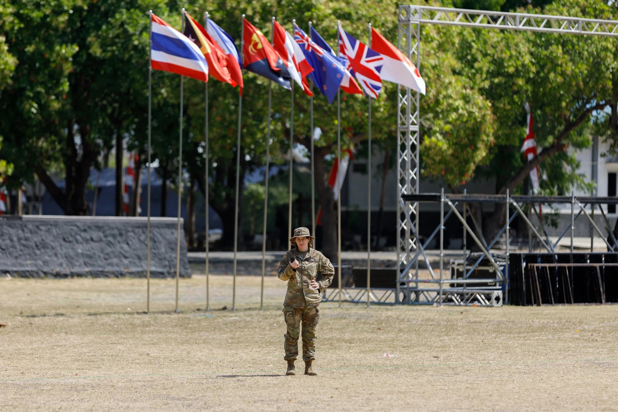 A soldier standing on a grassy field, holding a walkie-talkie, with multiple flags on poles and metal scaffolding in the background.
