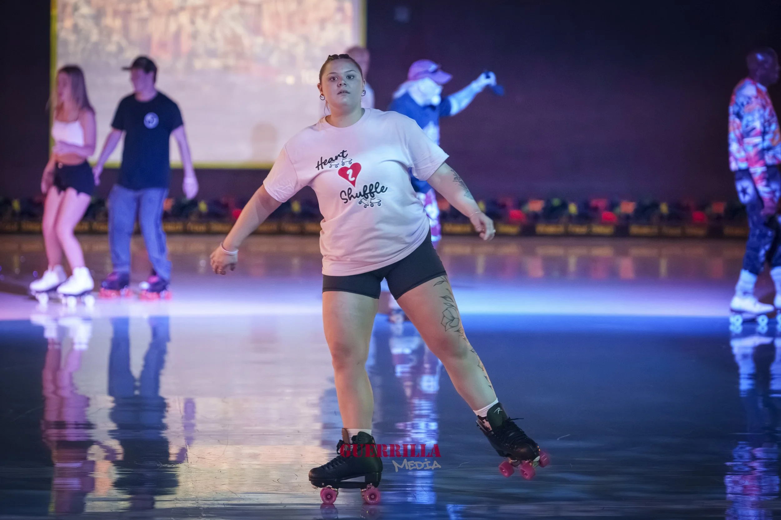 A woman roller skating on an indoor rink with colorful lighting, wearing a white T-shirt, black shorts, and black skates, with other skaters in the background.