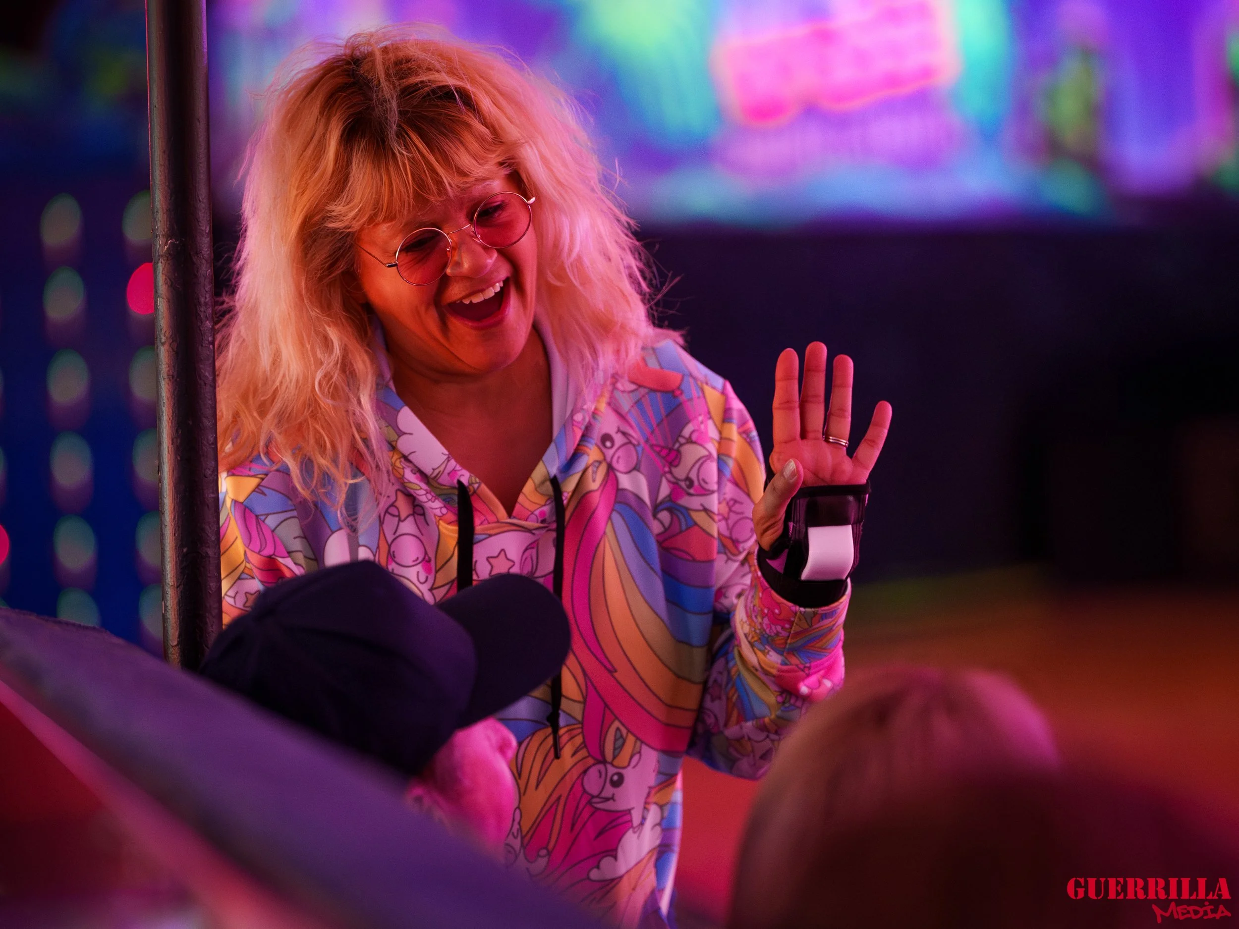 A woman with curly blonde hair wearing round glasses and a colorful, patterned top is smiling and waving at a person wearing a black cap in a setting with vibrant, neon lighting.