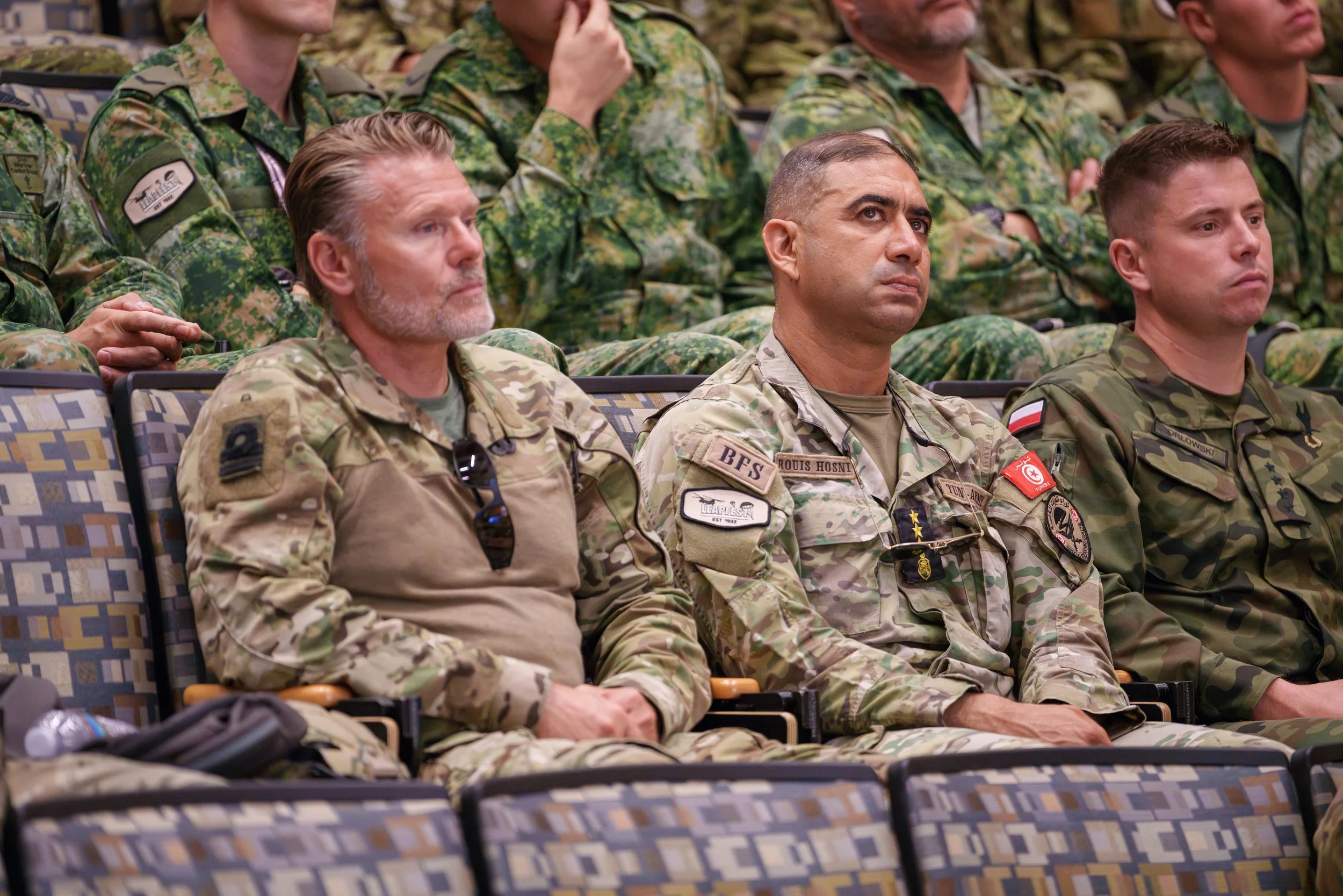 Military personnel sit in a row, attentively listening to a presentation or speech, wearing camouflage uniforms in an indoor setting.