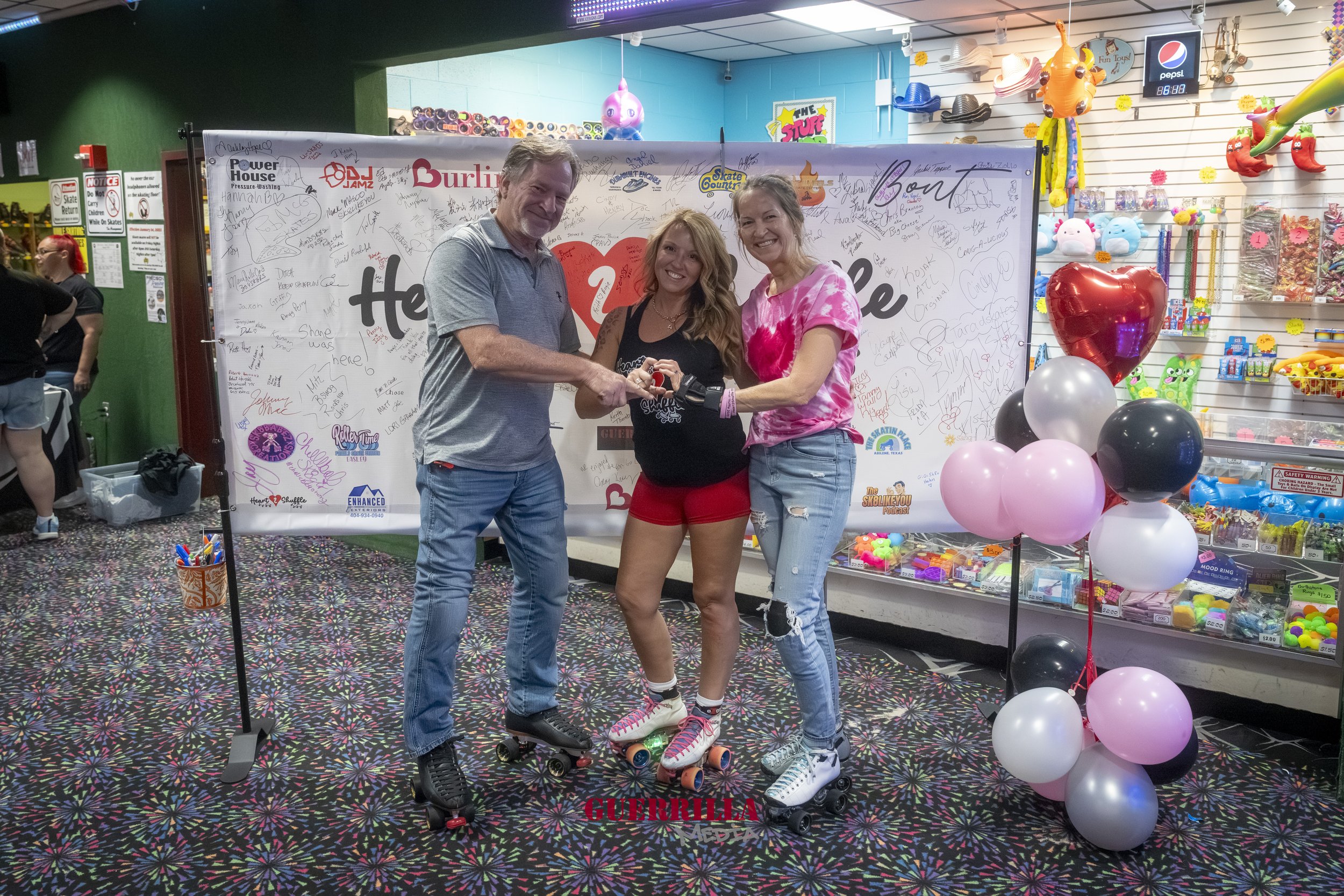 Three people on roller skates holding a key in front of a backdrop with signatures and logos, with balloons and toys on display in the background.
