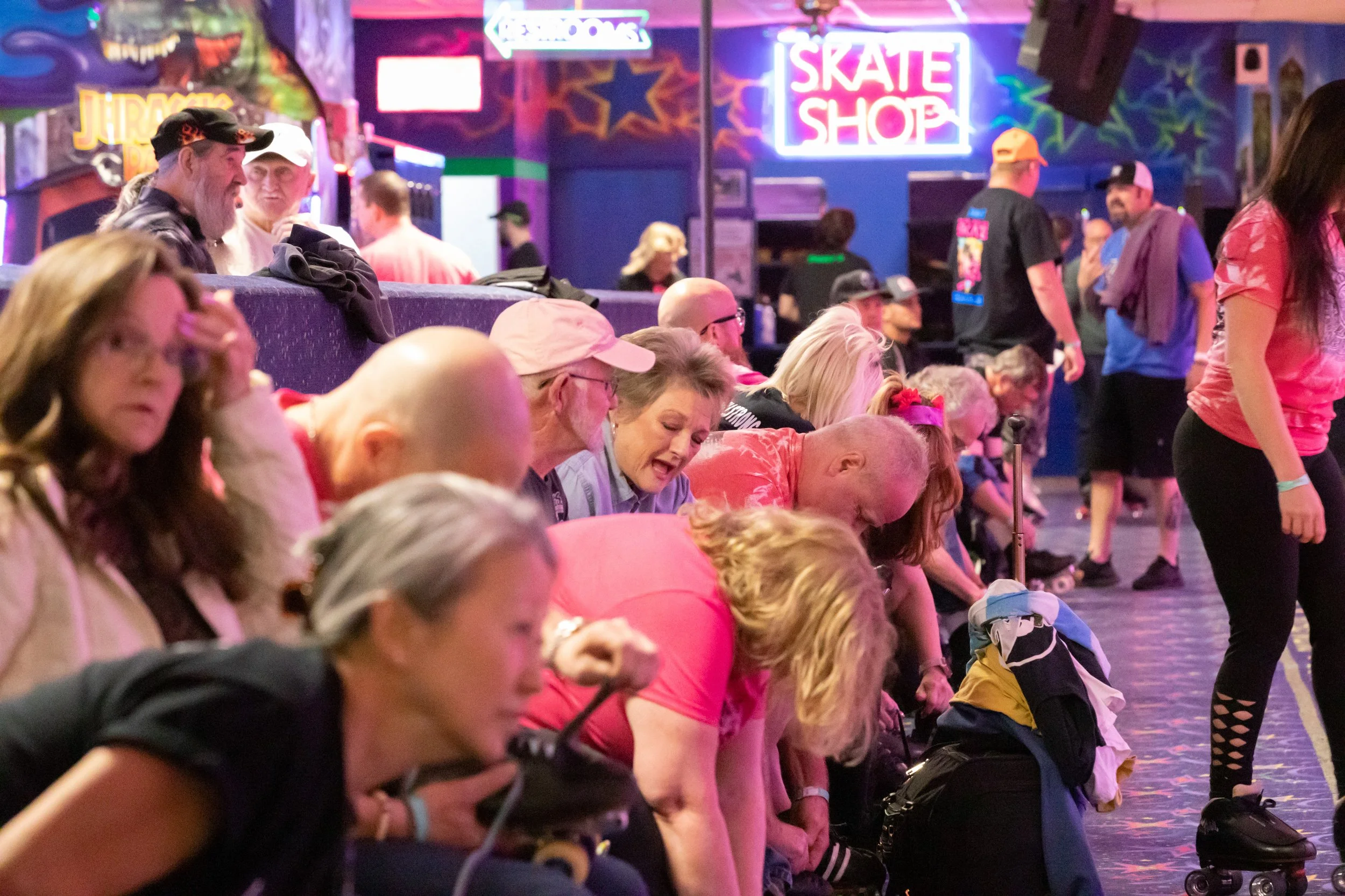 People at a roller skating rink, some sitting on the floor and others standing, with neon signs and colorful murals in the background.