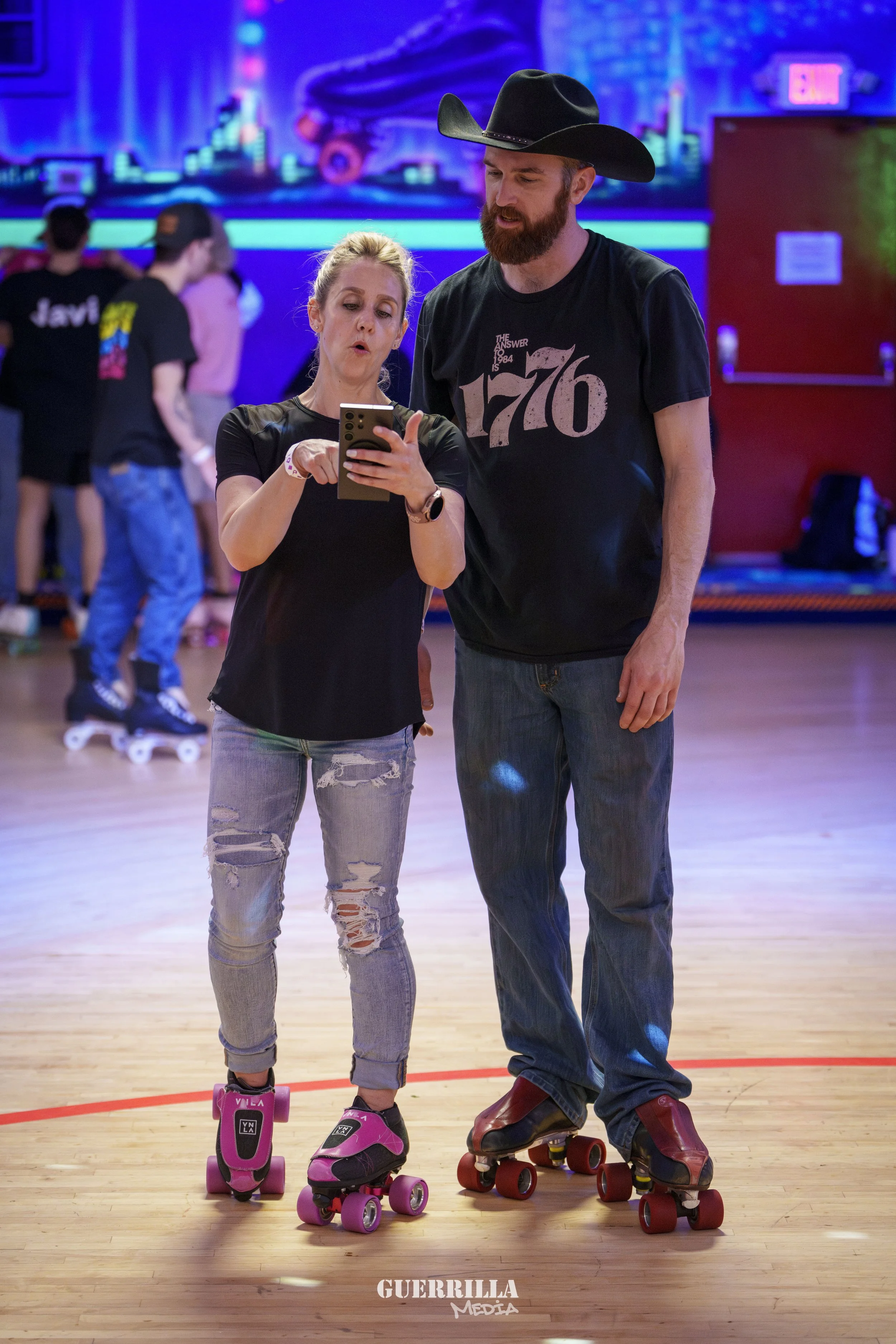 A woman and a man wearing roller skates, standing on a roller rink, looking at a smartphone together, with a colorful neon-lit background and other skaters in the distance.