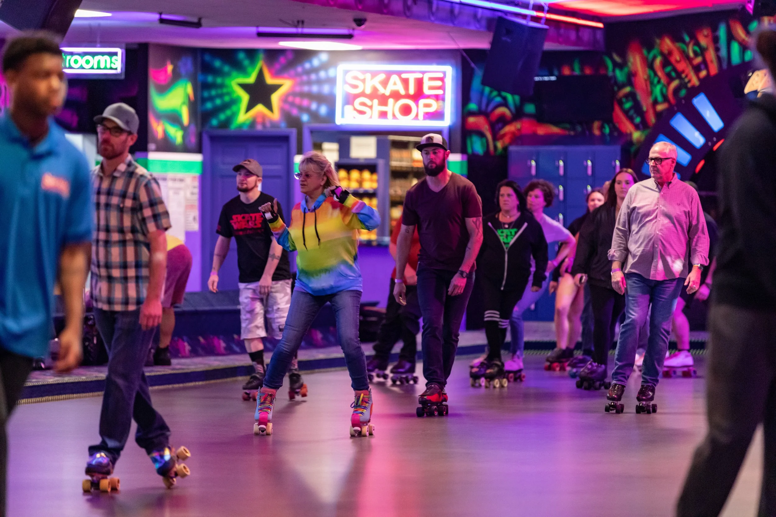 People roller skating inside a vibrant, neon-lit roller rink with colorful walls and signs, including a visible 'Skate Shop' sign.