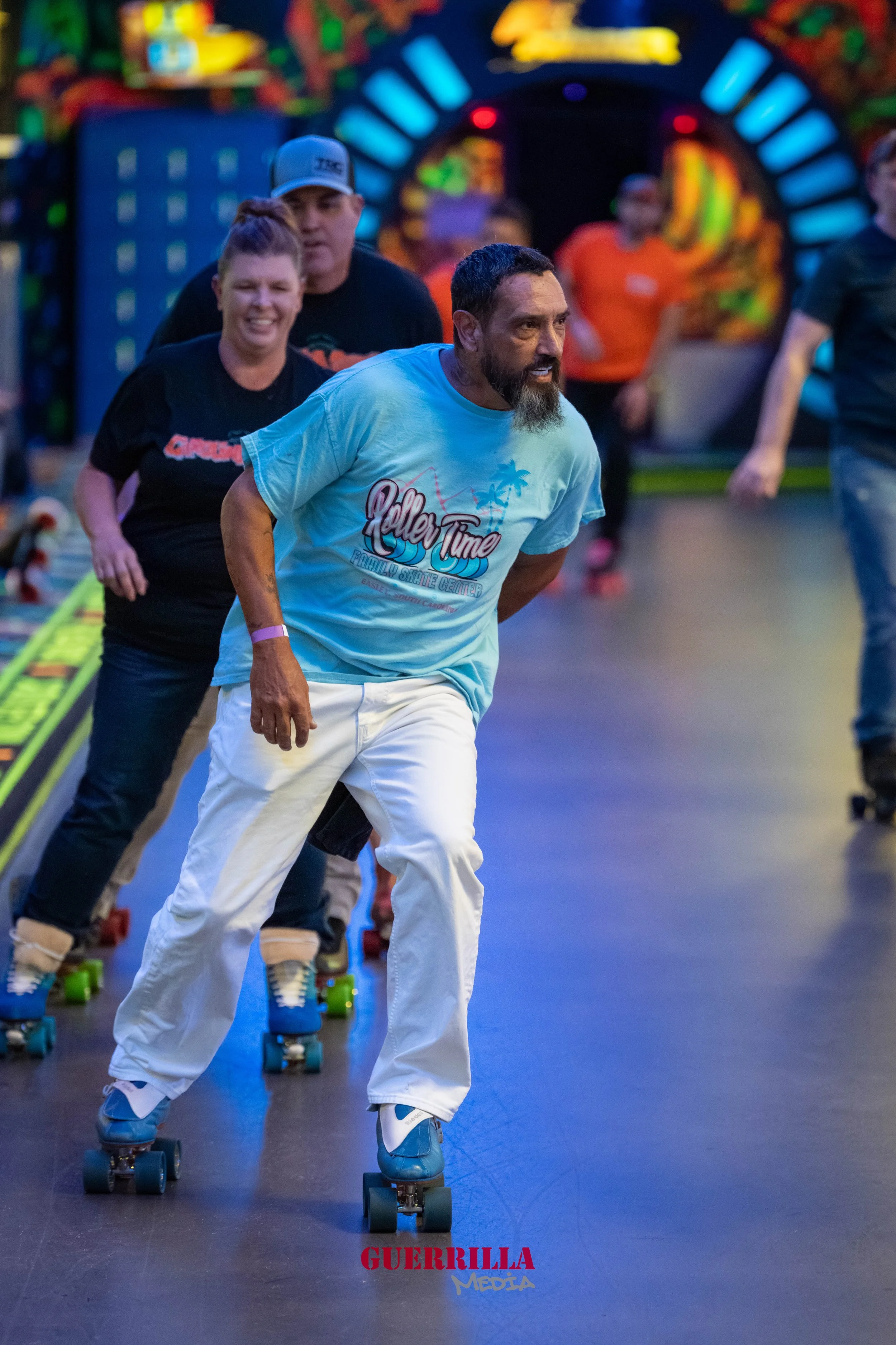 A group of people roller skating indoors with a colorful, neon-lit background. The man in the foreground is wearing a light blue t-shirt with a palm tree design and white pants, and appears to be leading or demonstrating skating. Behind him, a woman 