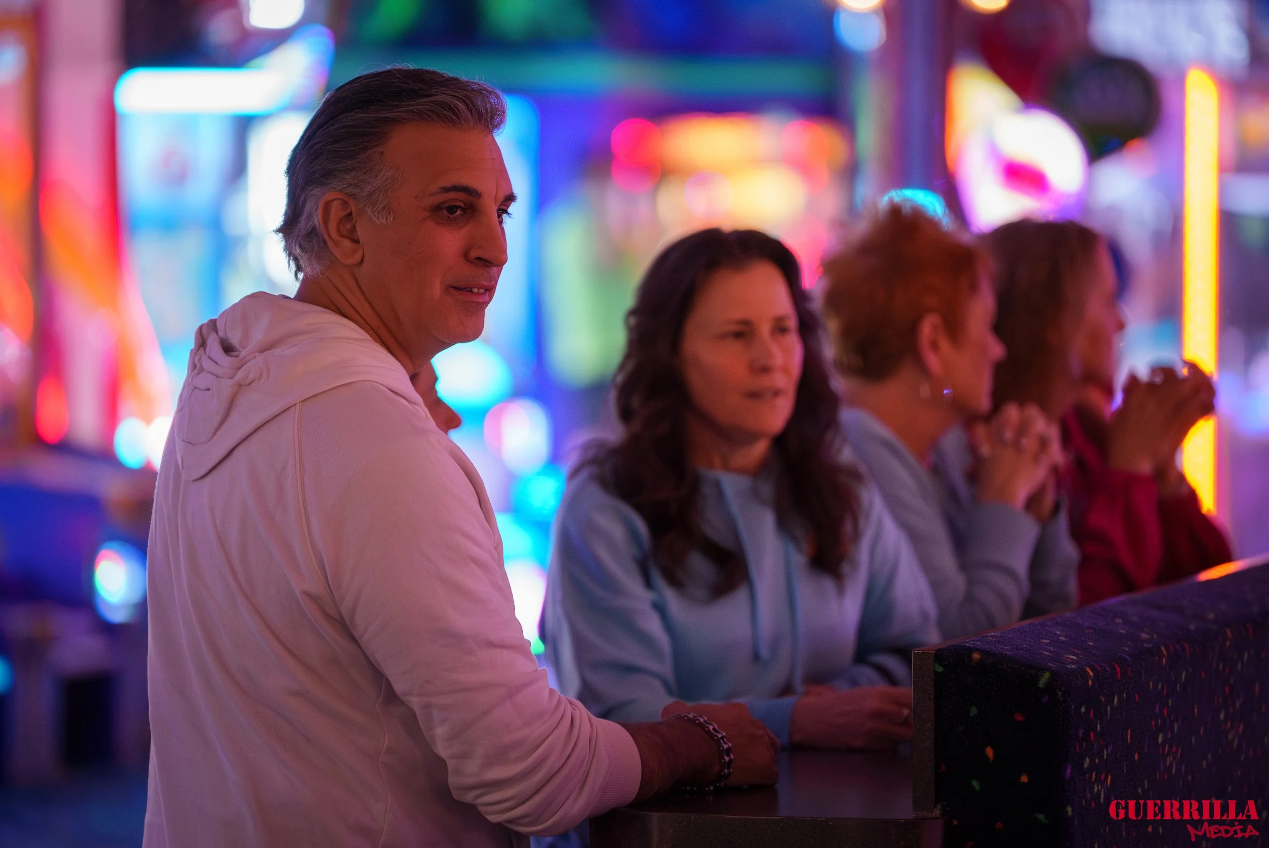 Four women standing at a counter in front of colorful bright lights at a carnival or fair, with the woman on the left looking at the camera.