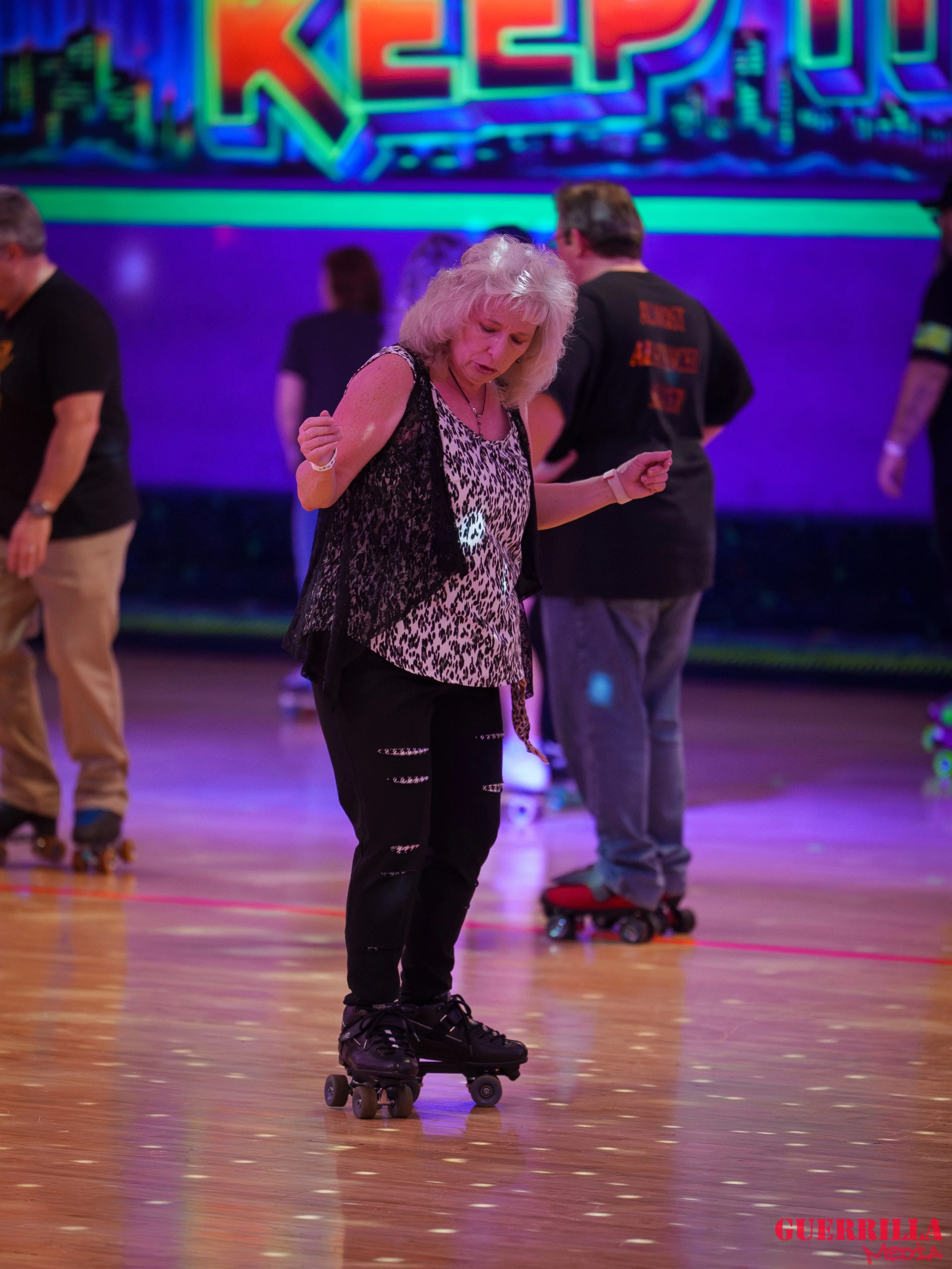 A woman with gray hair roller skating on a wooden floor at a rink, with other skaters and a neon sign in the background.