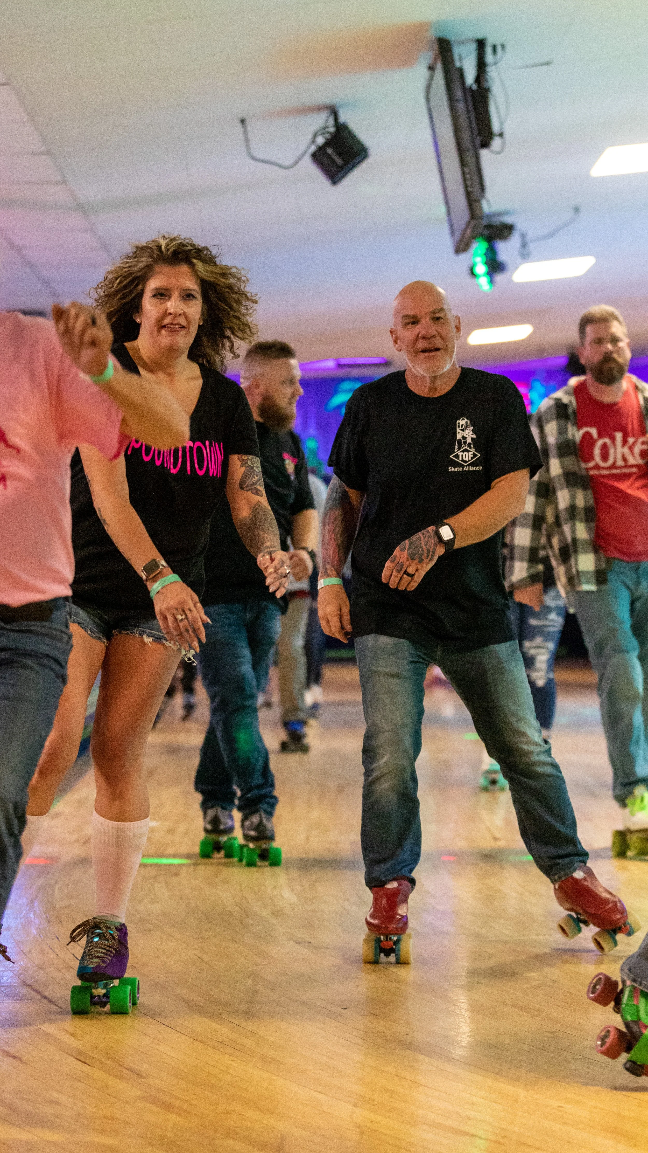 People roller skating indoors, some wearing casual clothes like t-shirts and jeans, enjoying a social skate session with colorful lights and a wooden floor.