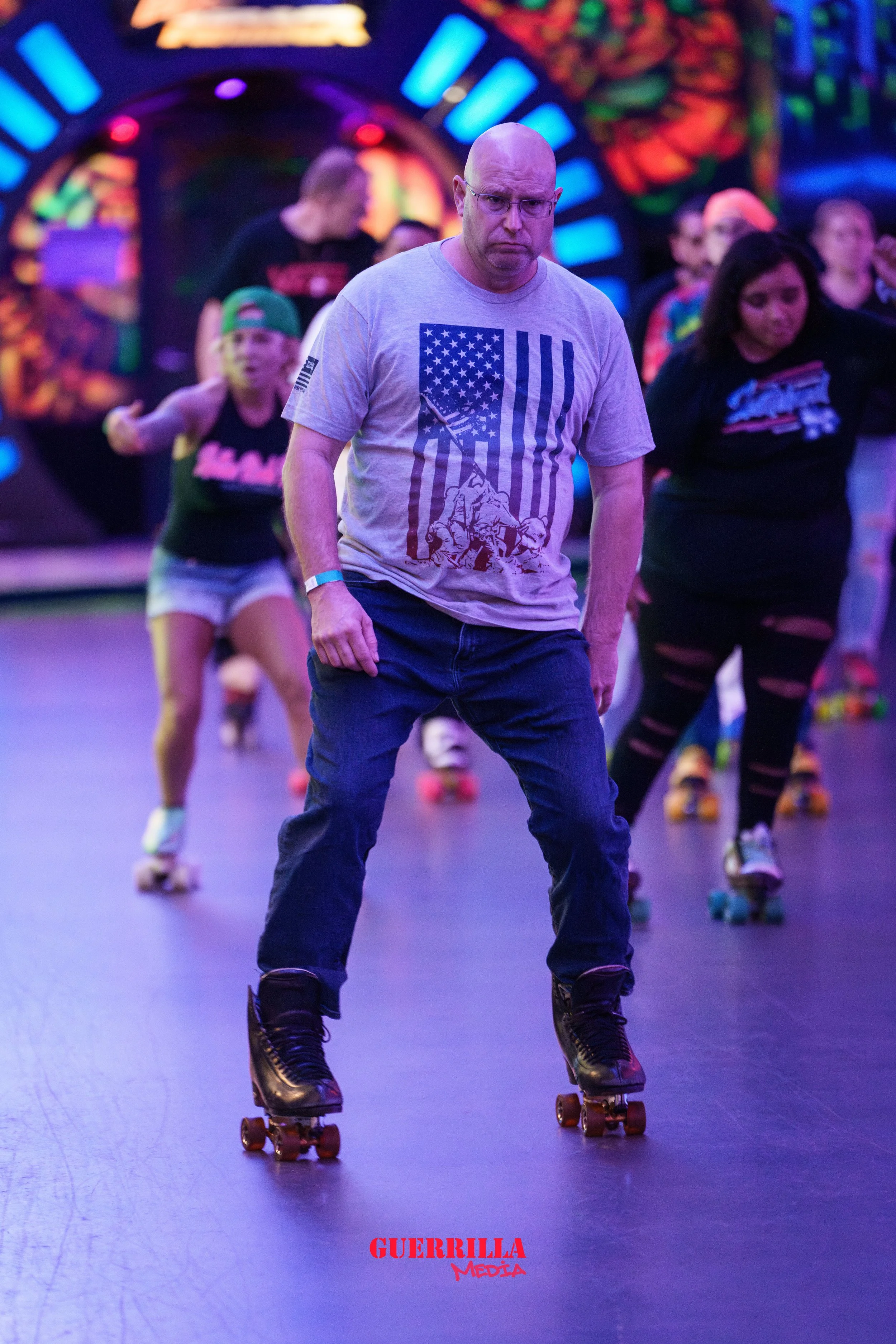 A man wearing glasses and a gray T-shirt with an American flag and a soldier graphic, skating at an indoor roller skating rink with colorful neon lights, with a girl in a black tank top and jean shorts and a woman in a black ripped T-shirt and black 