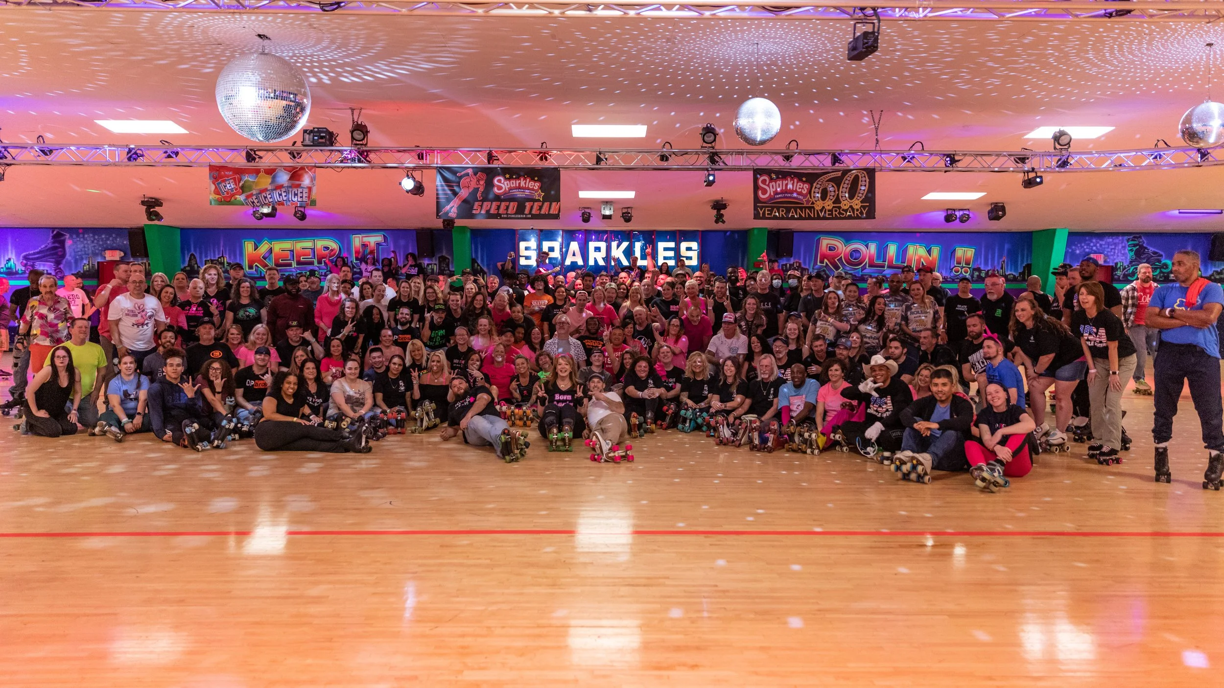 A large group of people gathered in a roller skating rink for a group photo. The background features neon signs with words like 'Keep It', 'Sparkles', and 'Rollin', and disco balls hang from the ceiling.