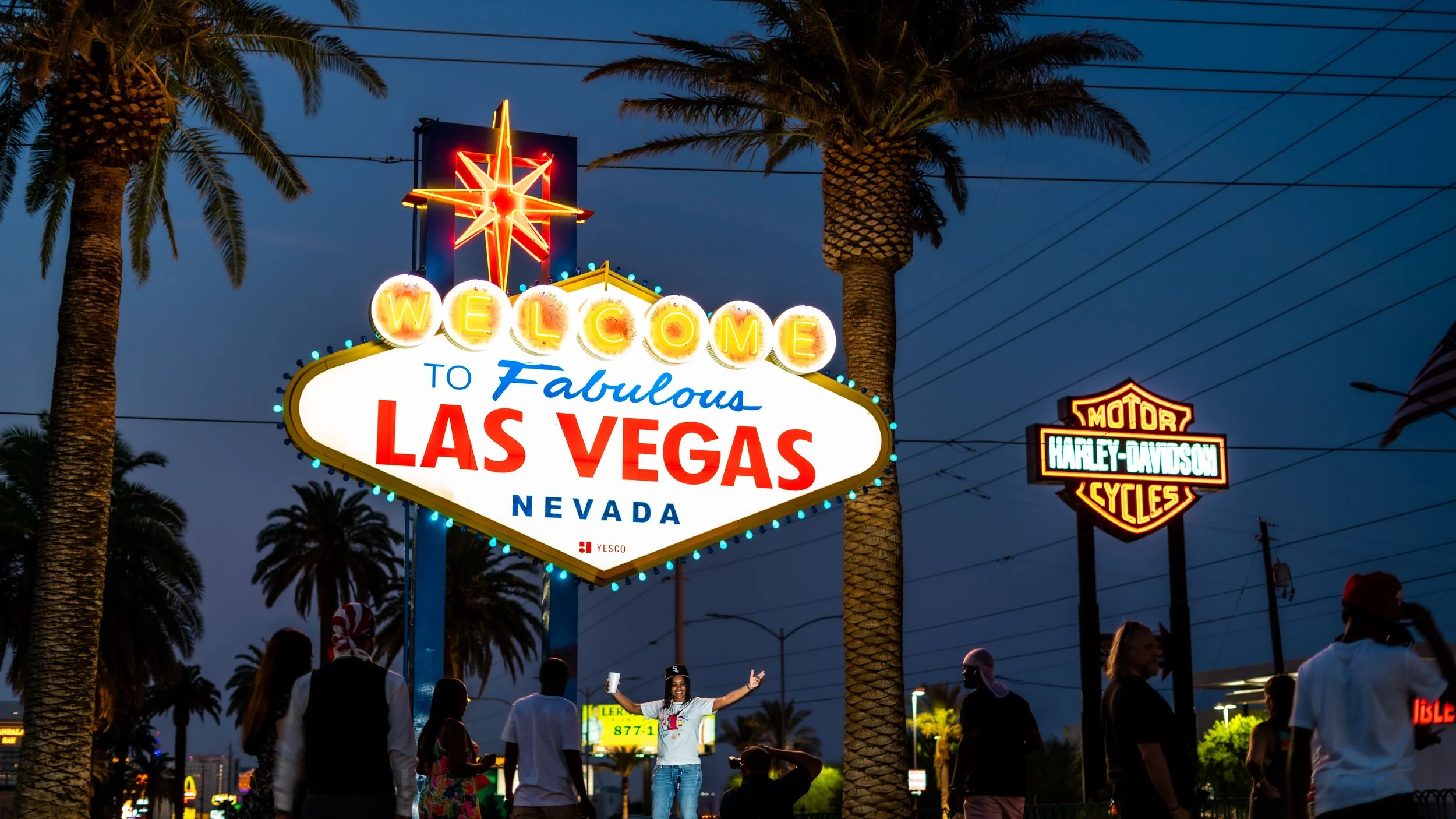 Nighttime scene at the Las Vegas Welcome sign, featuring illuminated neon letters and a group of people, including a woman in a white shirt taking photos, palm trees, and Harley-Davidson signs in the background.