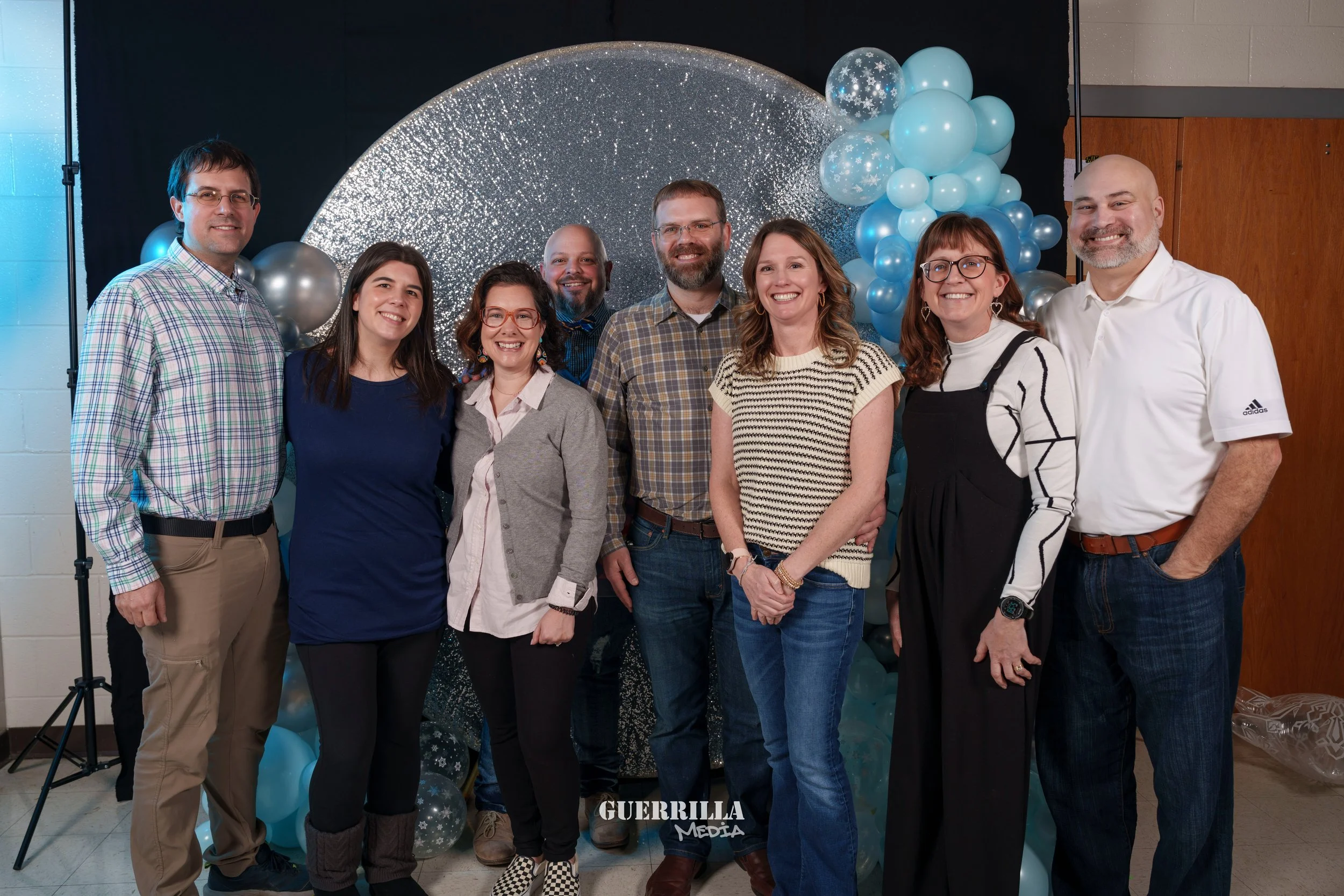 Group of nine people standing in front of a sparkly blue and silver balloon backdrop for a celebration.