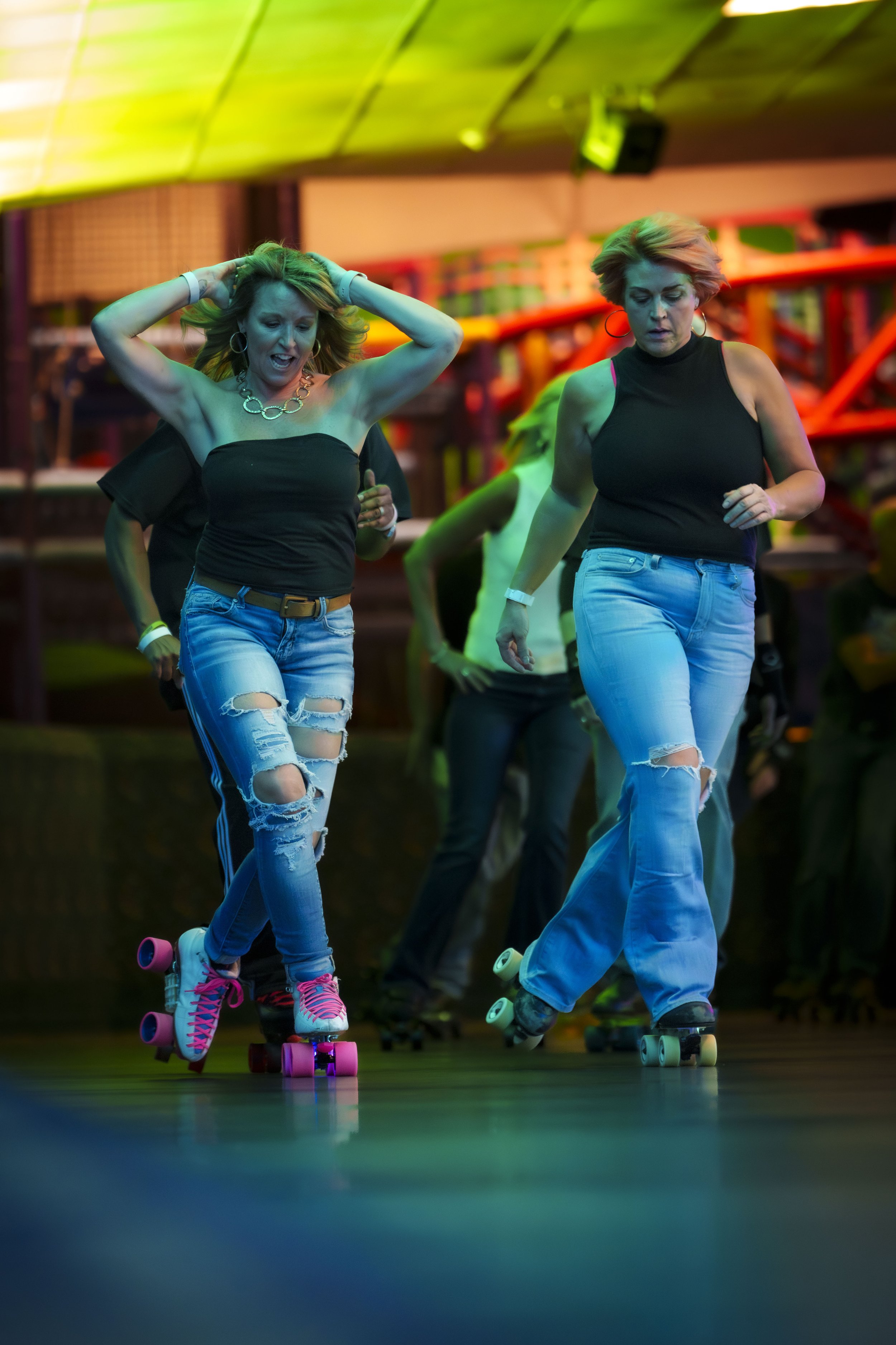 Two women roller skating indoors, wearing ripped jeans and black tops, with colorful neon lighting and a lively environment.