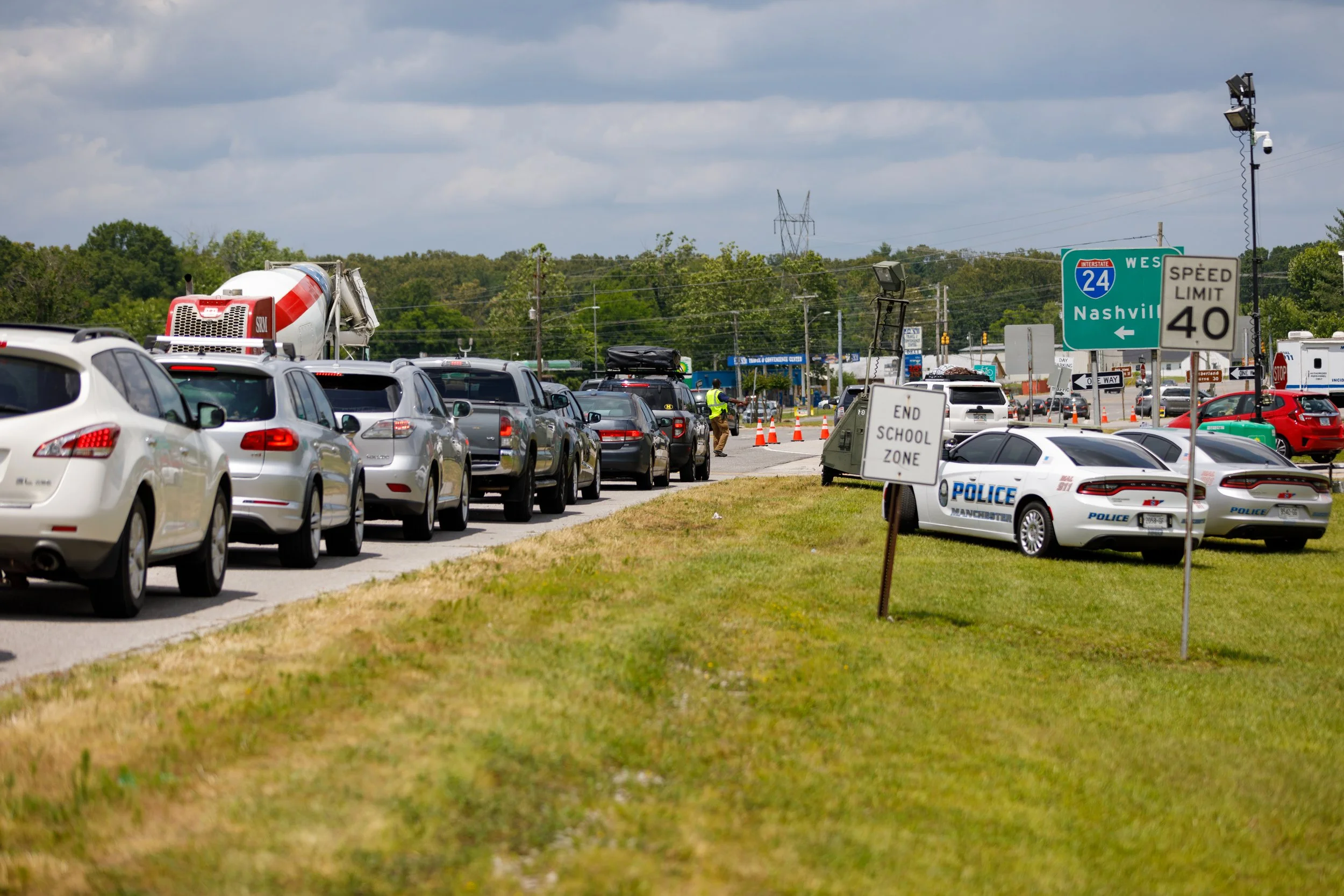Traffic jam with line of cars near a school, police cars, and school zone signs, with traffic cones and police officers managing the scene, around a sign indicating end of school zone and speed limit.