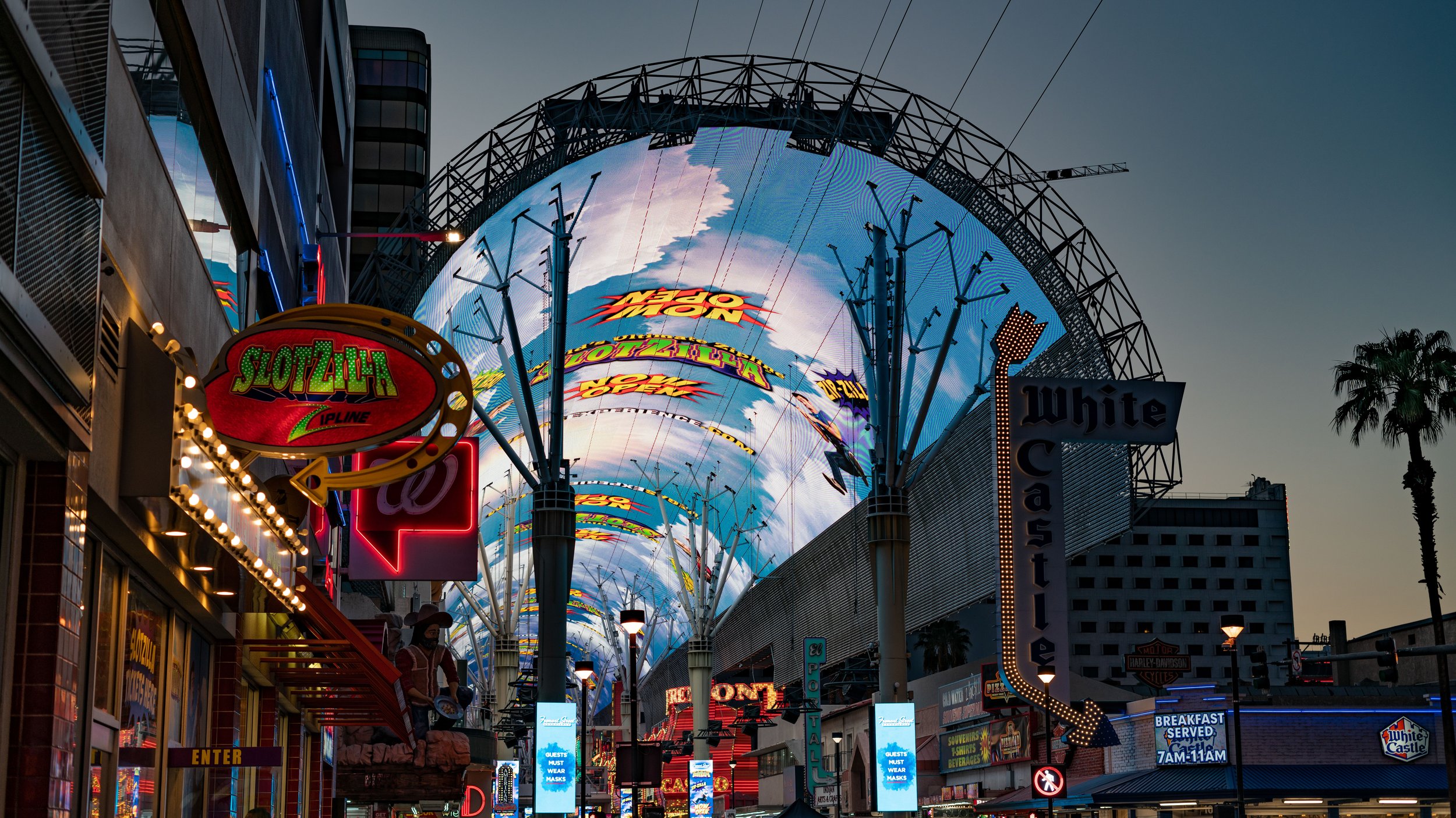Nighttime view of the Las Vegas Strip with illuminated signs including SlotZilla and White Castle, large LED digital billboards, street lights, palm trees, and various storefronts.