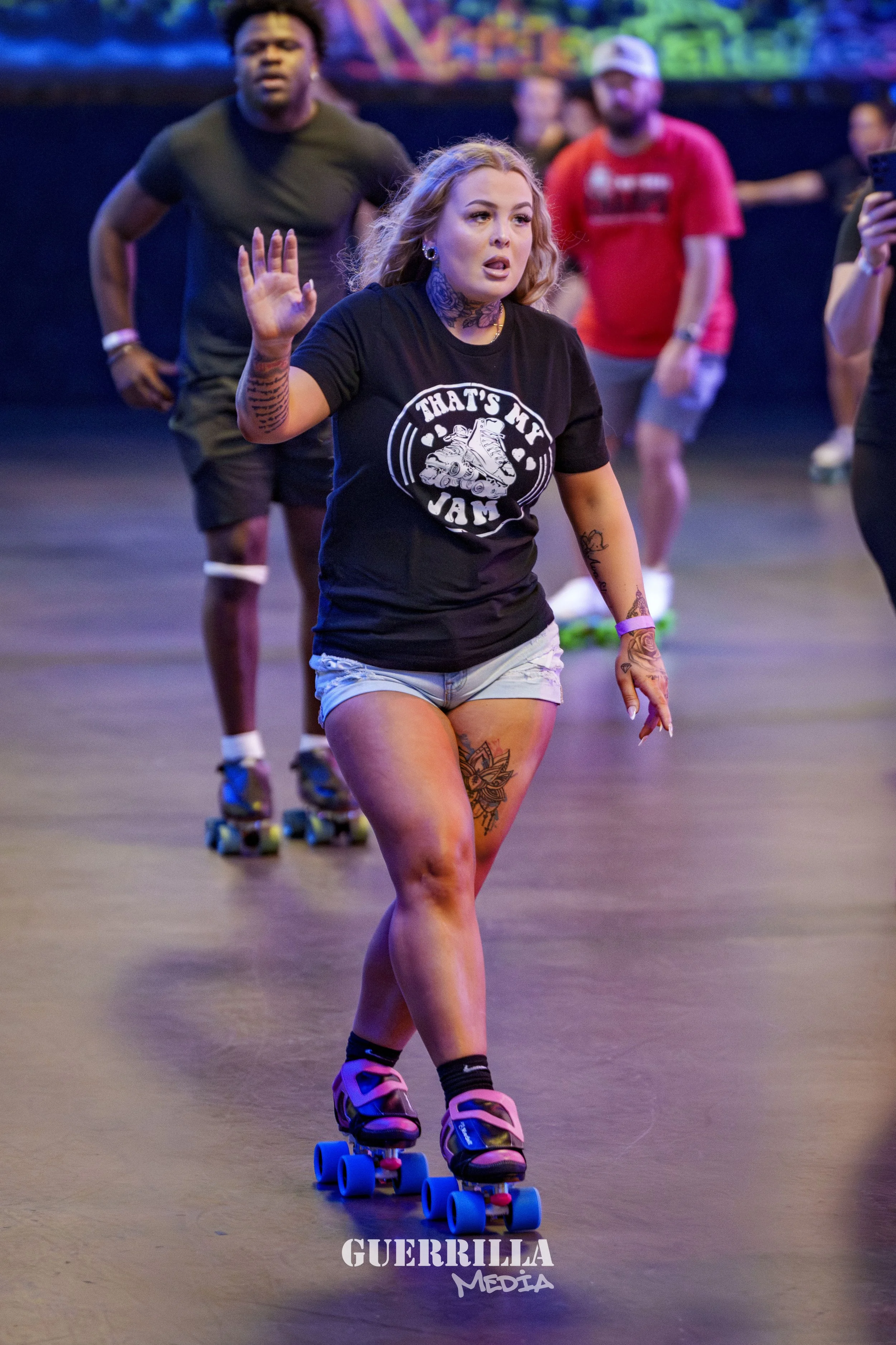 A woman roller skating indoors, wearing a black T-shirt with 'That's My Jam' and a graphic of a sneaker, and light-colored shorts. She has tattoos on her arms and legs and is holding her hand up as she skates. Other people are in the background also 