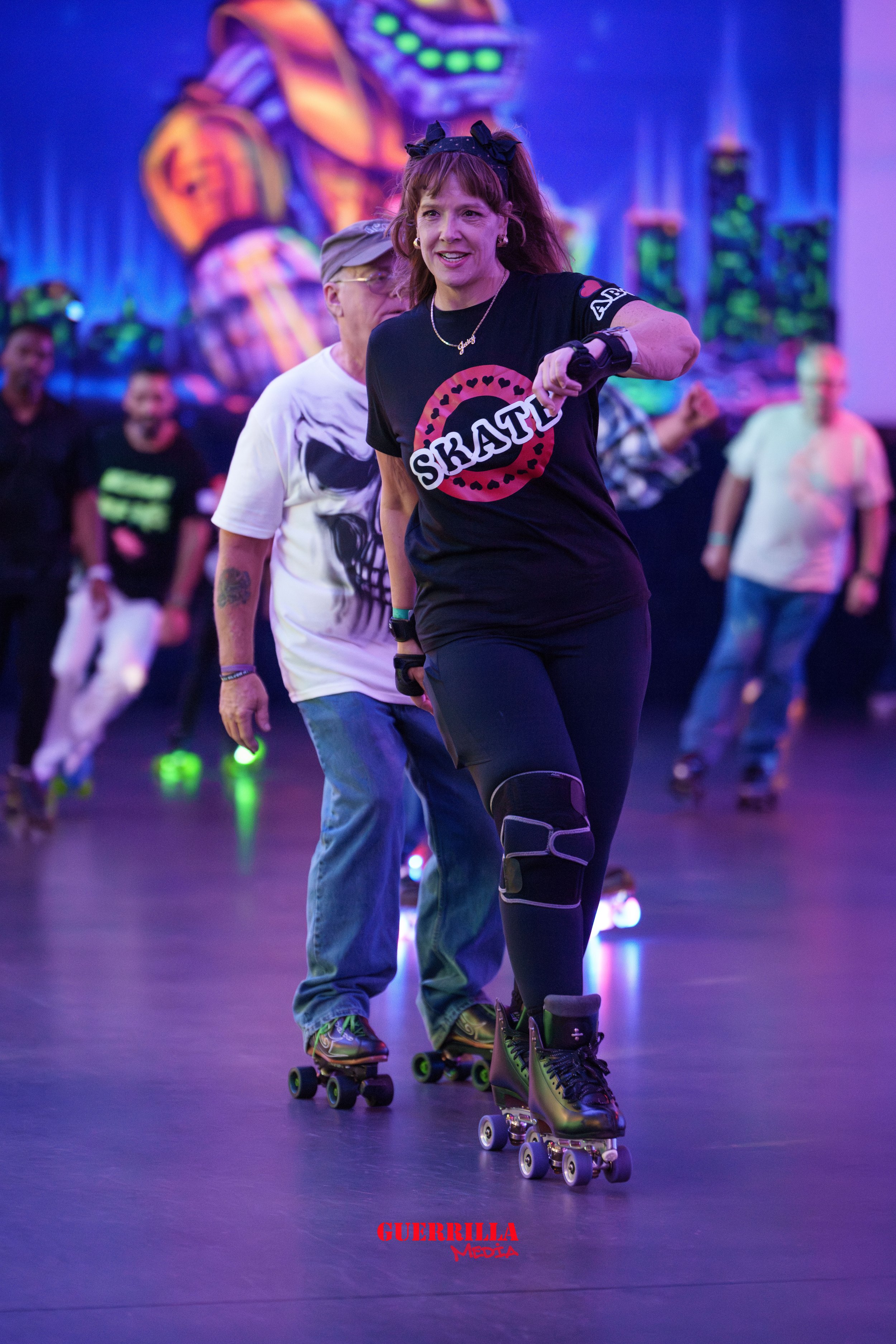 A woman roller skating in a roller rink with neon lighting, wearing a black t-shirt with sketch and 'SKATE' text, black leggings, and headband, with others roller skating in the background.