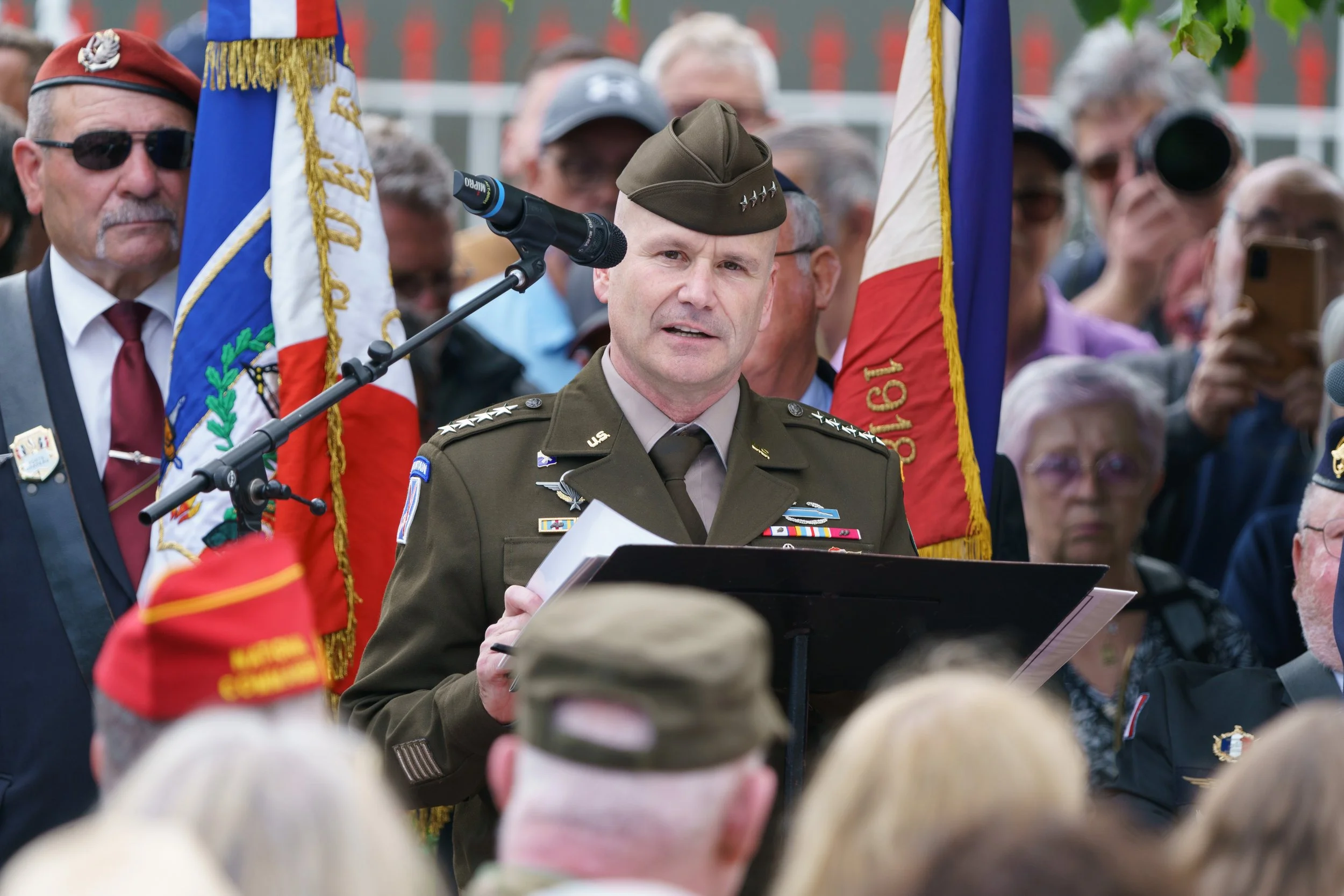 A man in a U.S. military uniform speaking at a podium during a ceremony, with Veterans flags and a crowd of people in the background.