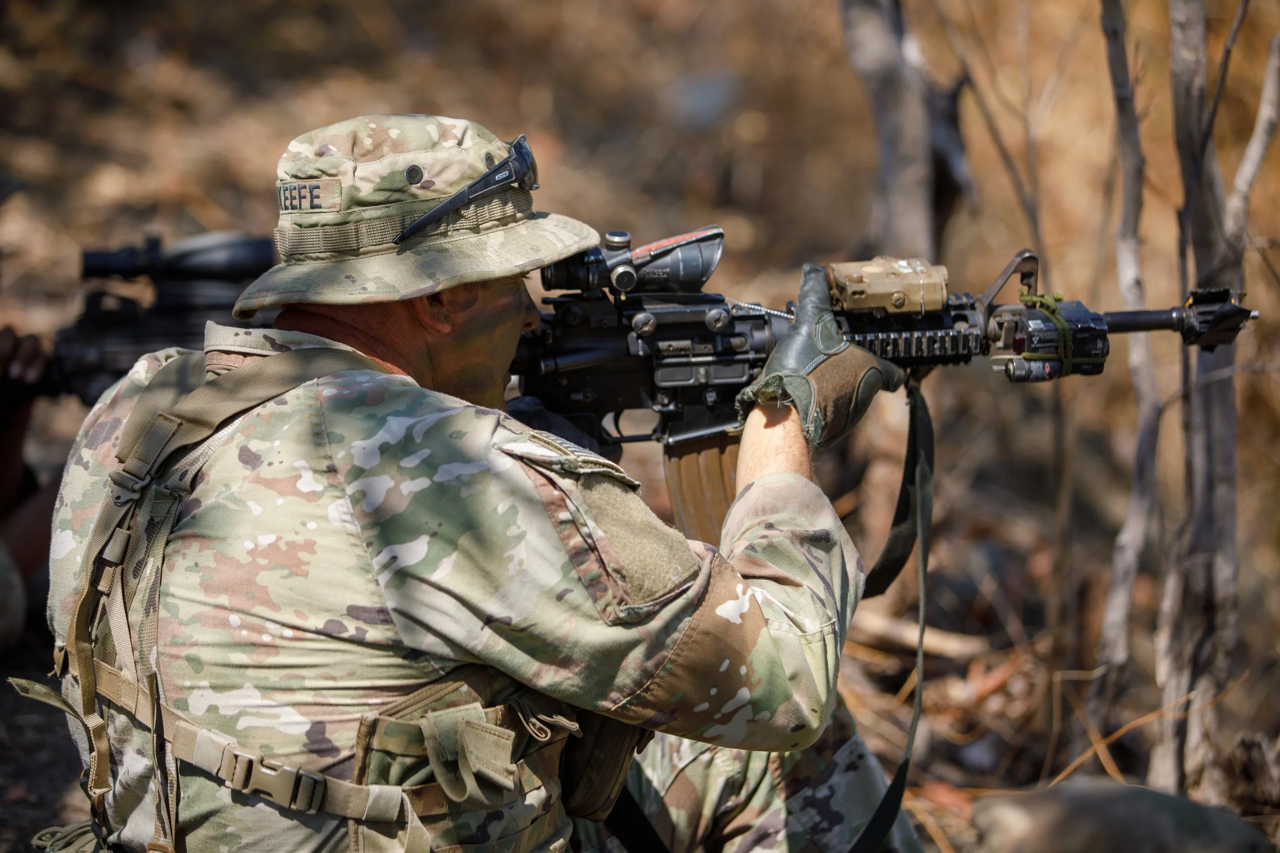 A soldier in camouflage uniform aiming a rifle in a wooded outdoor environment.