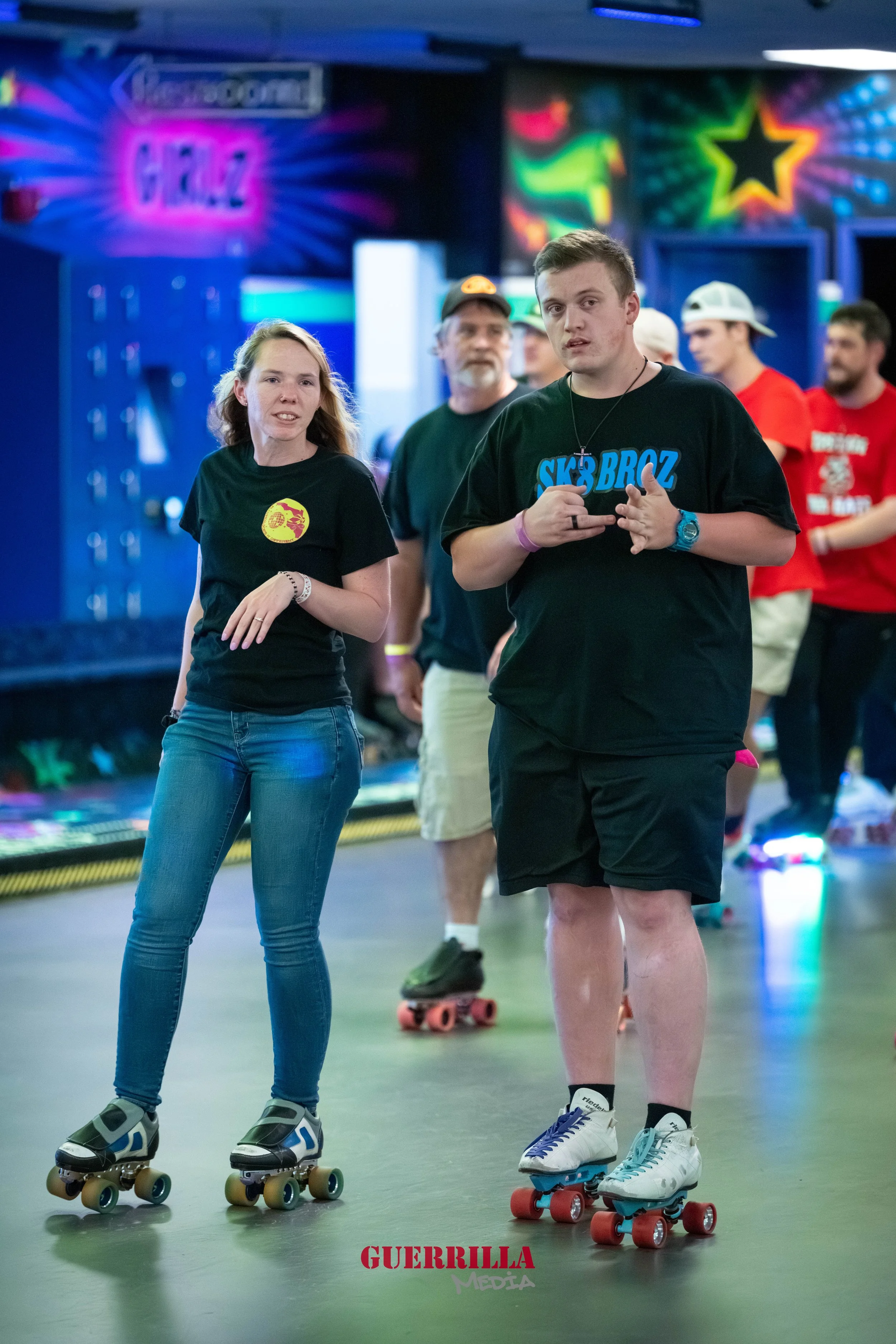 People roller skating at an indoor rink with colorful neon wall art and signs in the background, including the word 'GUERRILLA' at the bottom.