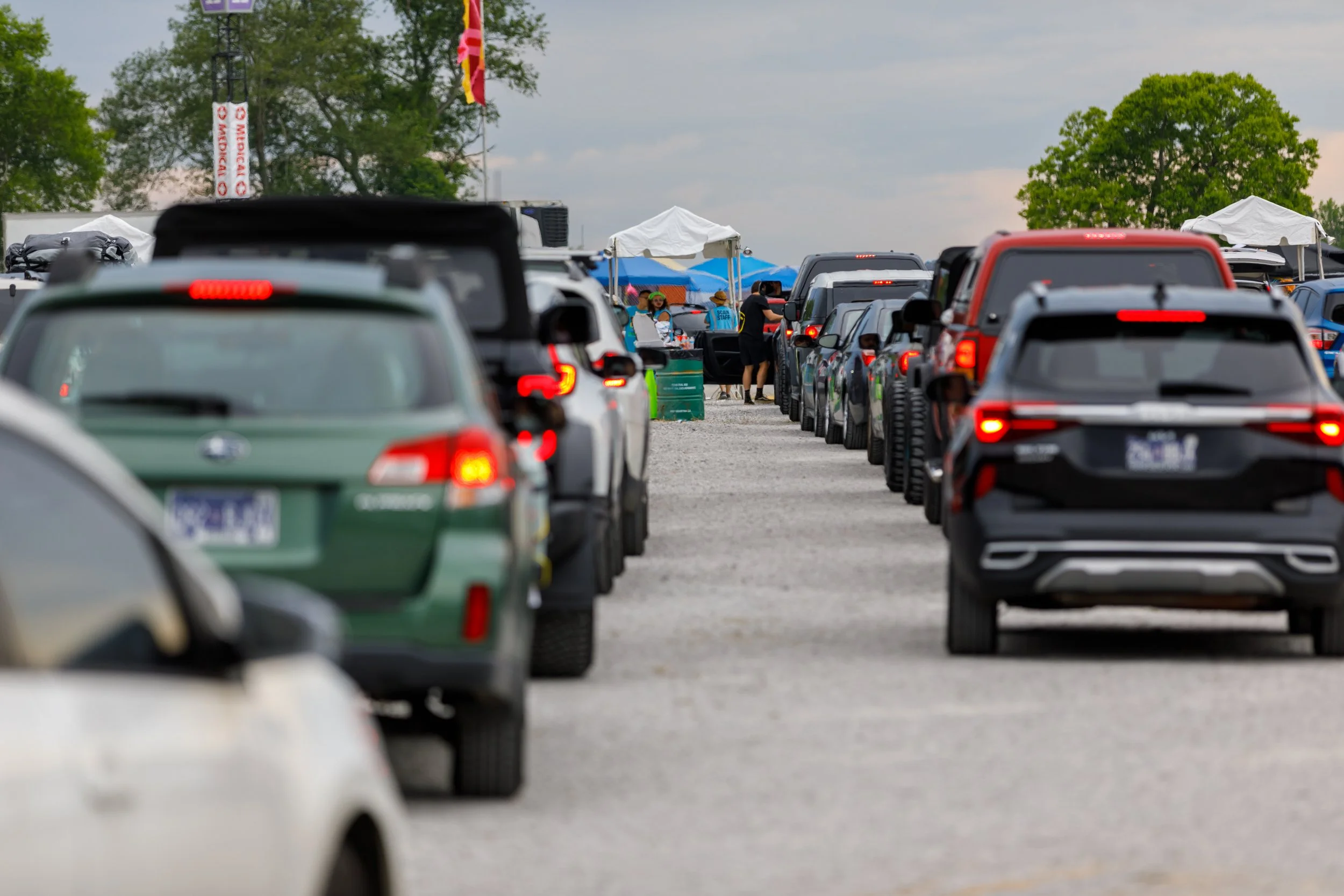 A long line of cars parked on a gravel surface at an outdoor event, with tents and people visible in the background.