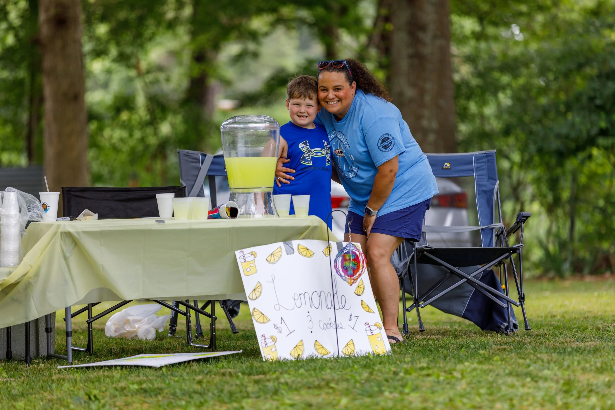 A woman and a young boy selling lemonade at a stand in a park with trees in the background. The sign indicates the lemonade costs $1 and s'mores cost $3.