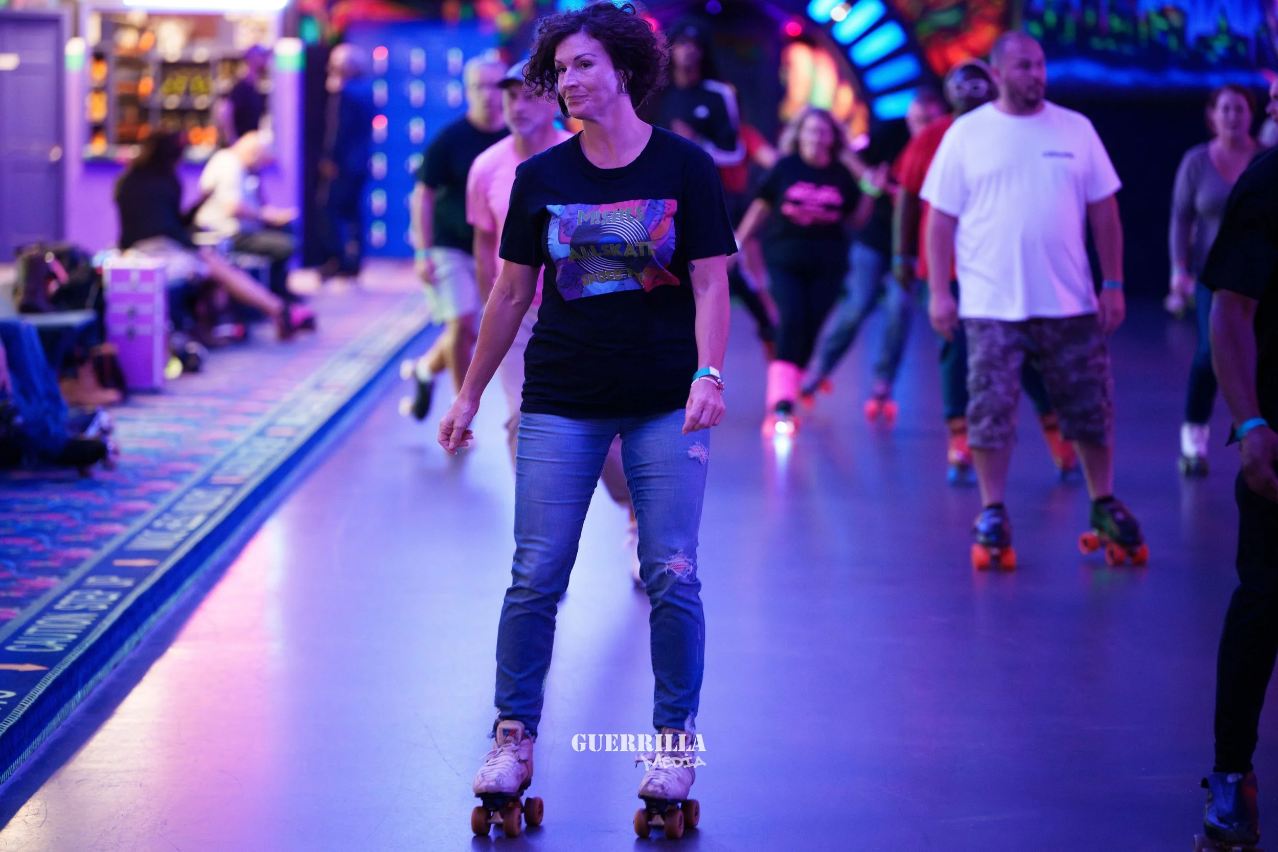 A woman roller skating indoors at a roller rink, surrounded by other skaters, with colorful neon lights in the background.