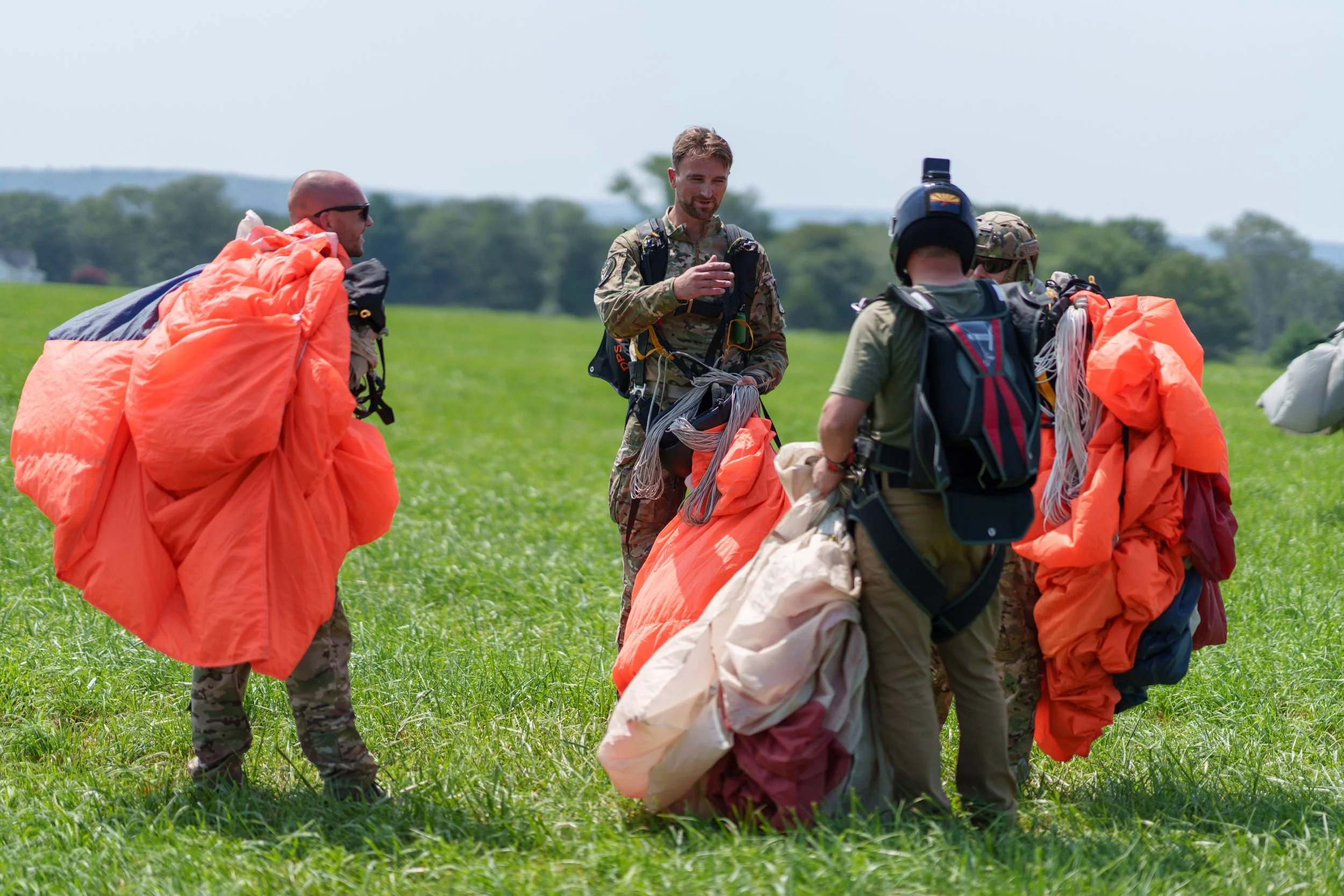 Group of skydivers dressed in military gear preparing for a jump in a grassy field.
